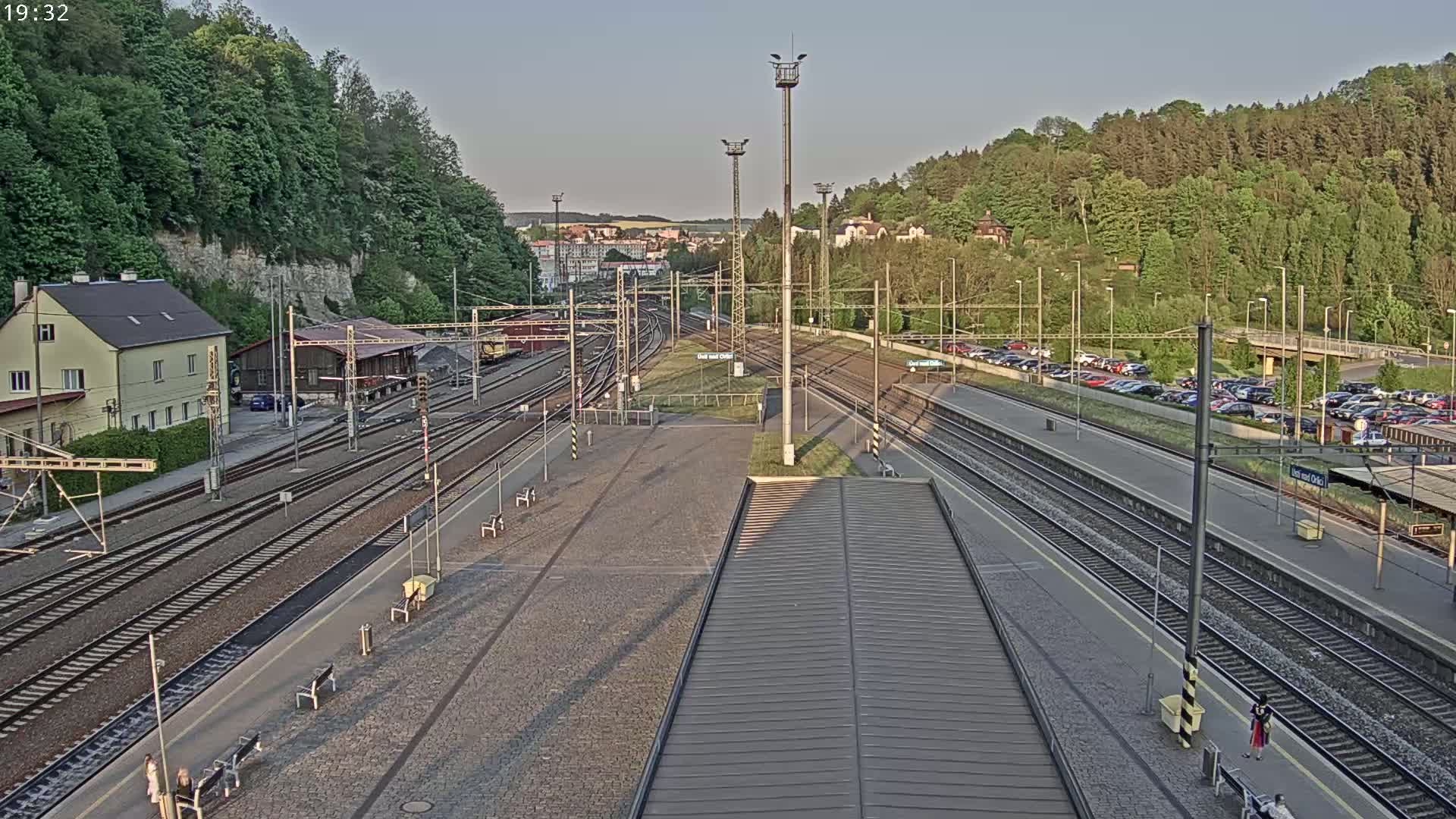 A high-angle, long shot of a train station at dusk, with multiple train tracks, a large parking lot, and a hill covered in lush green trees in the background.