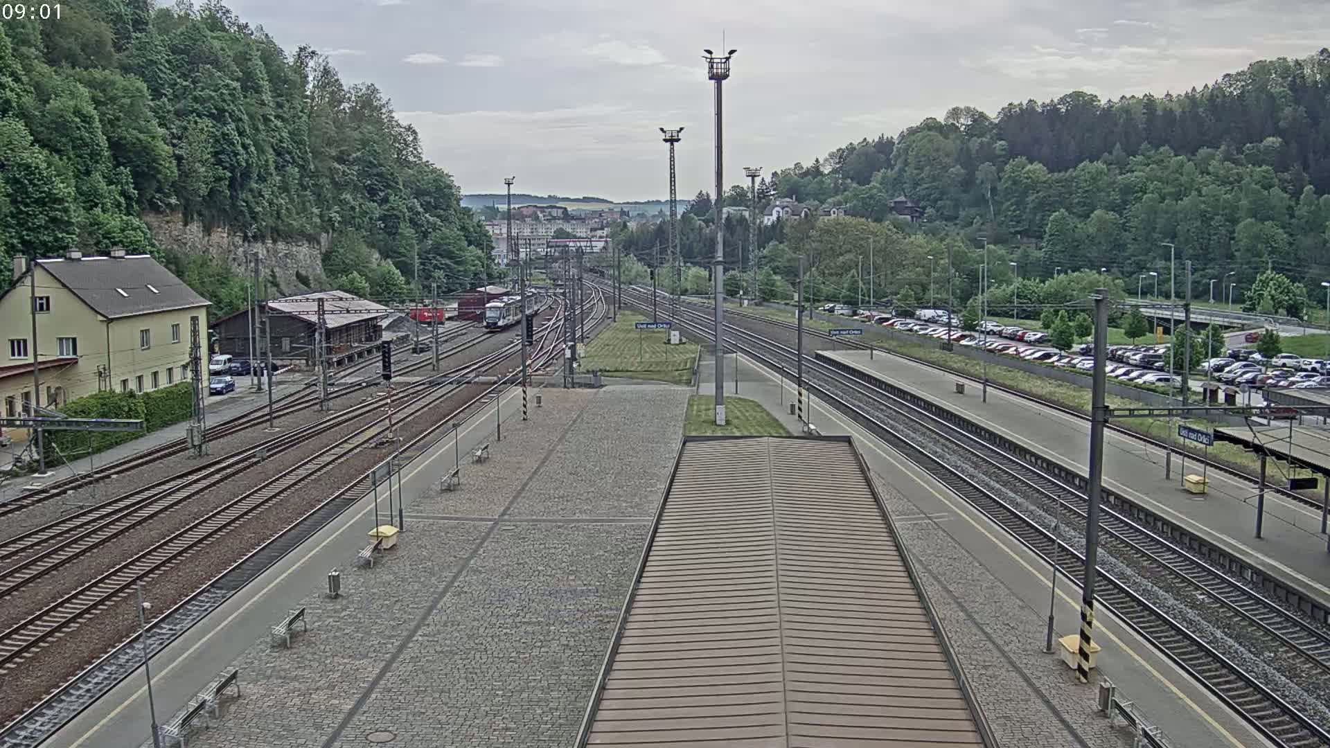 A train station with multiple train tracks, a building, and a parking lot is shown on an overcast day.