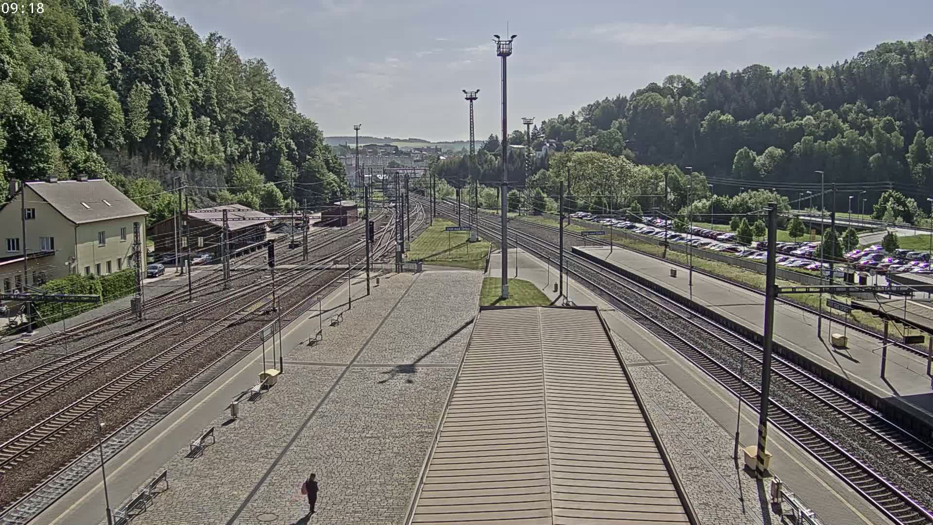 A mostly sunny view of a train station with multiple tracks, a large parking lot, and a person walking on the platform, all surrounded by lush green hills.