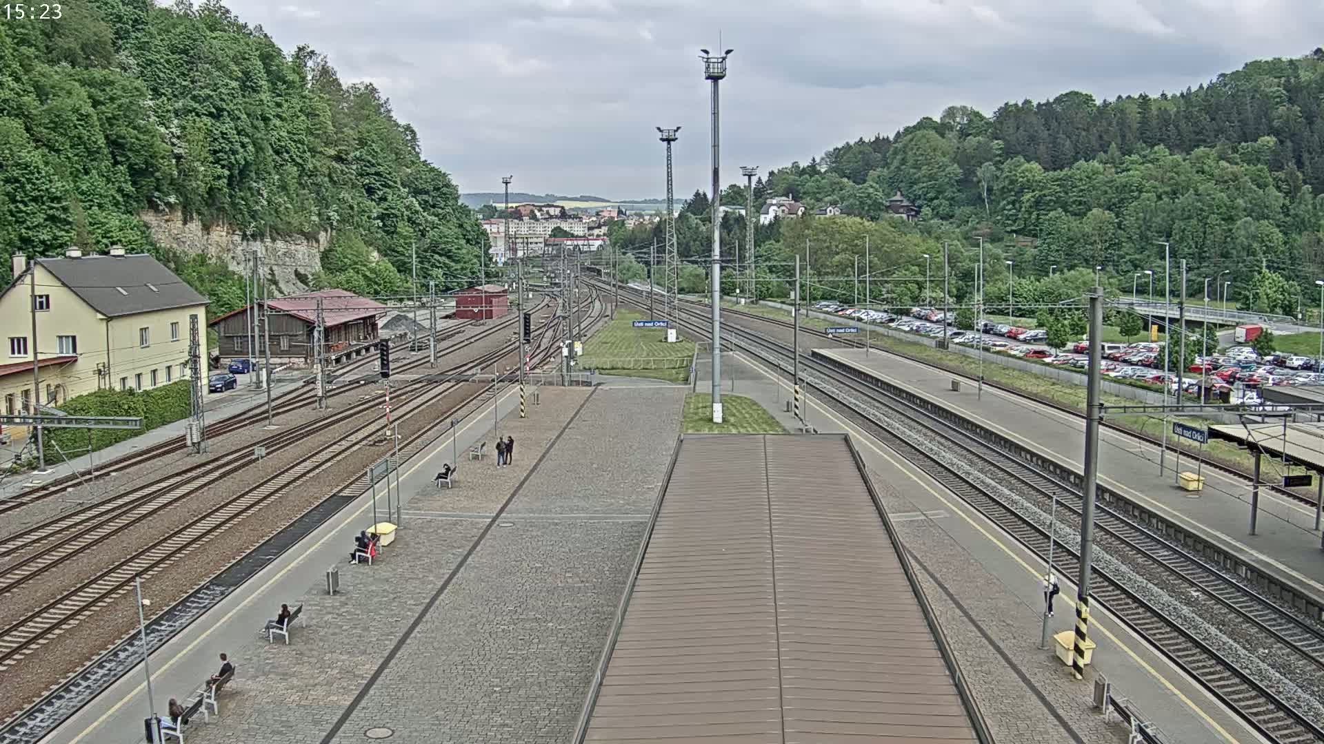 A train station with multiple tracks, platforms, and a parking lot is visible under an overcast sky, surrounded by lush green hills.