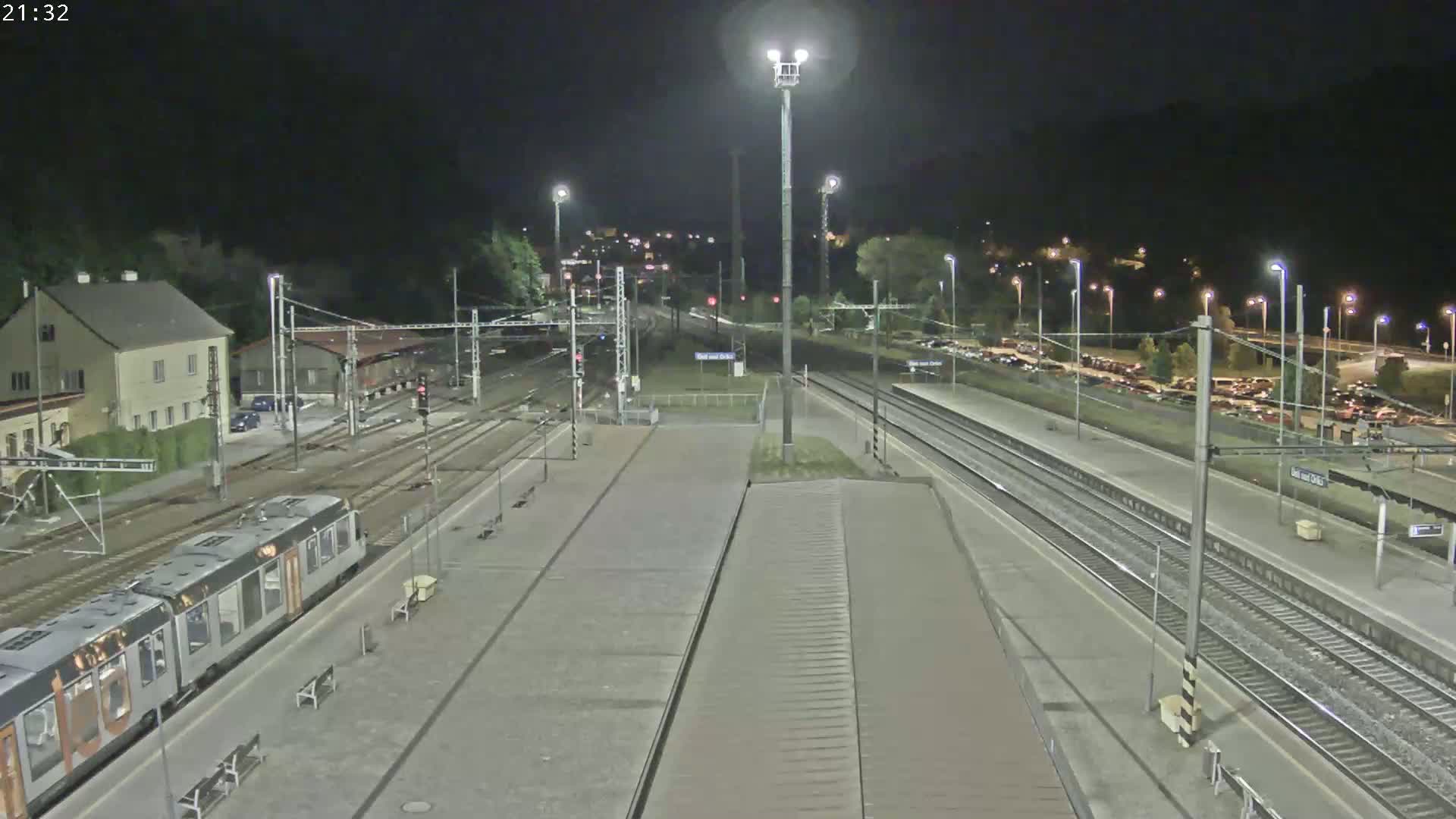 A nighttime view of a train station with a train on one of the platforms, surrounded by tracks, buildings, and a parking lot.
