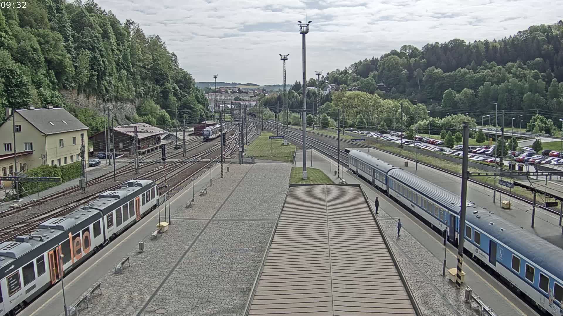 A train station with two trains on the platform, under an overcast sky, surrounded by green hills and a large parking lot.