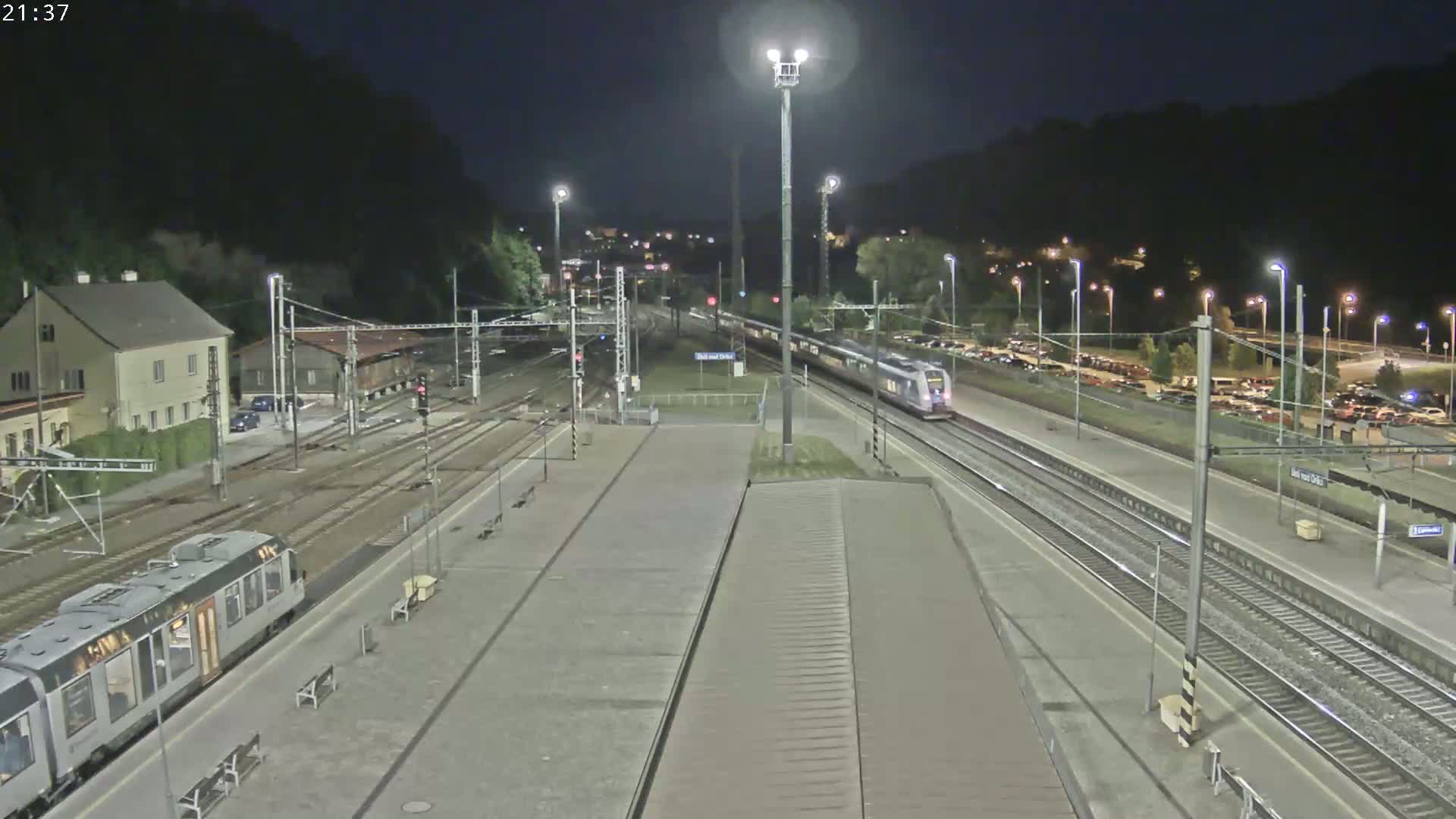 A nighttime view of a train station with a train passing through, and cars parked in a lot.