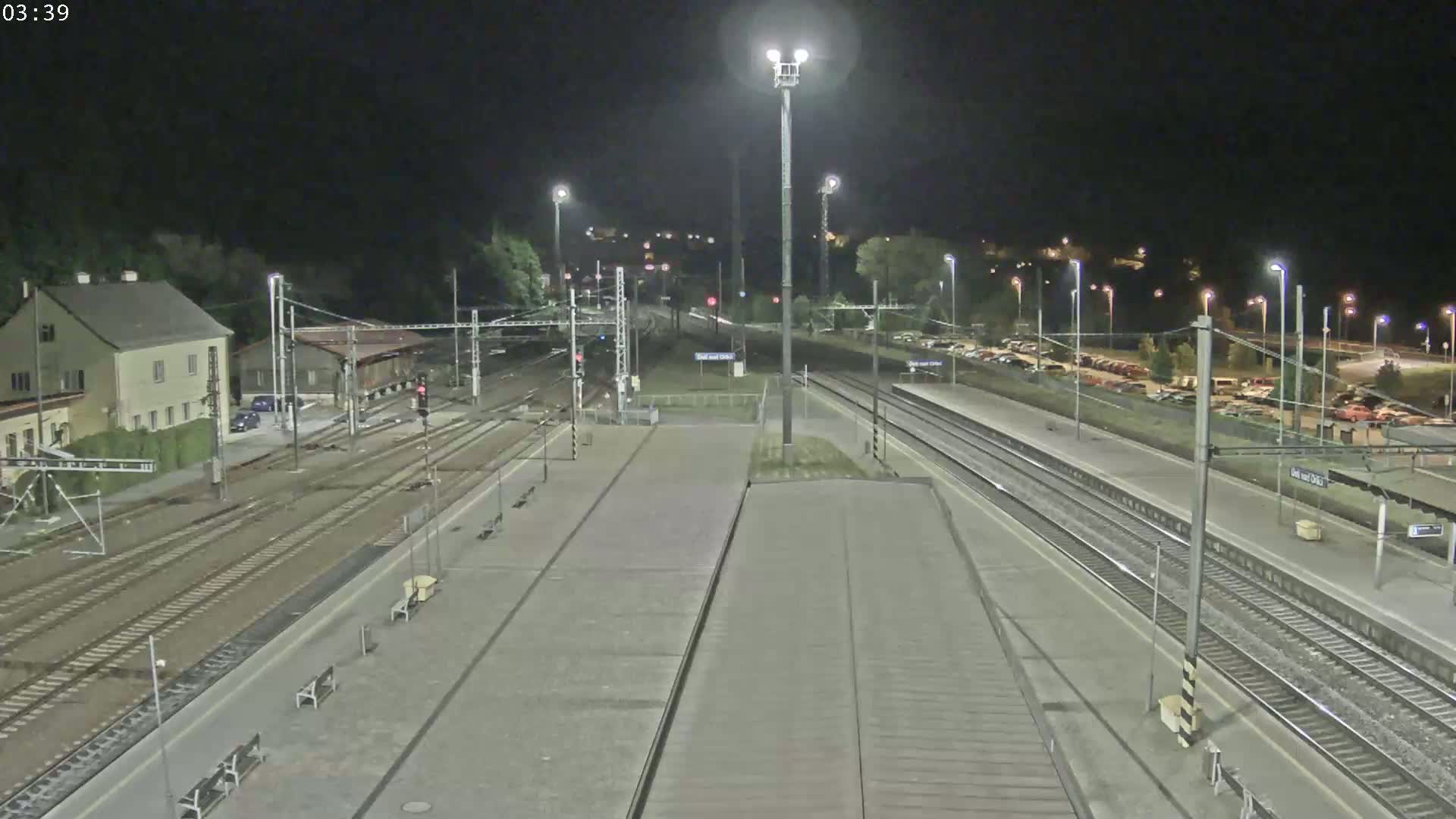 A nighttime view of a train station platform with several train tracks, a building, and a parking lot.