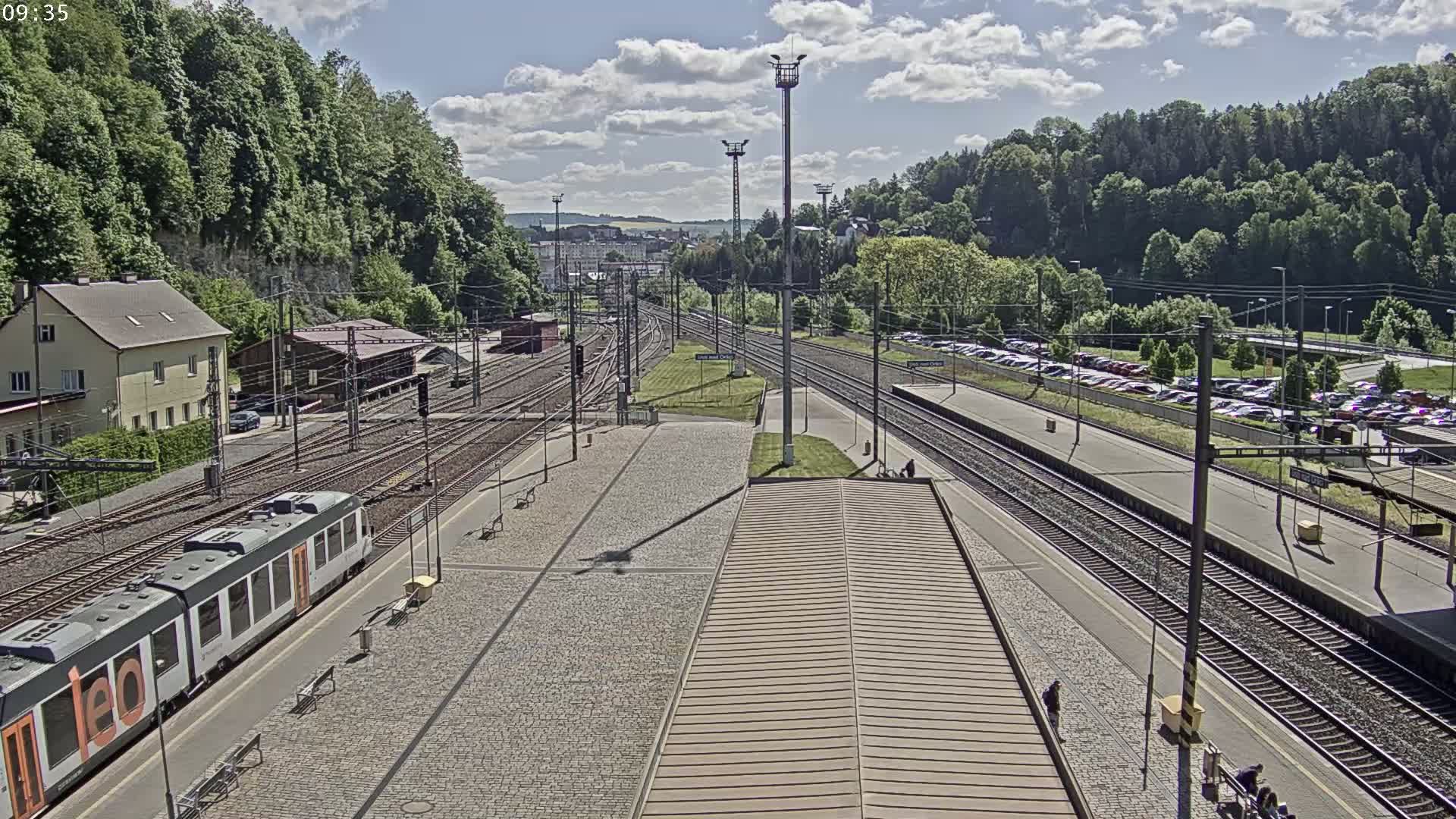 A train station on a sunny day with several train tracks, a passenger train, and a large parking lot visible in the background.