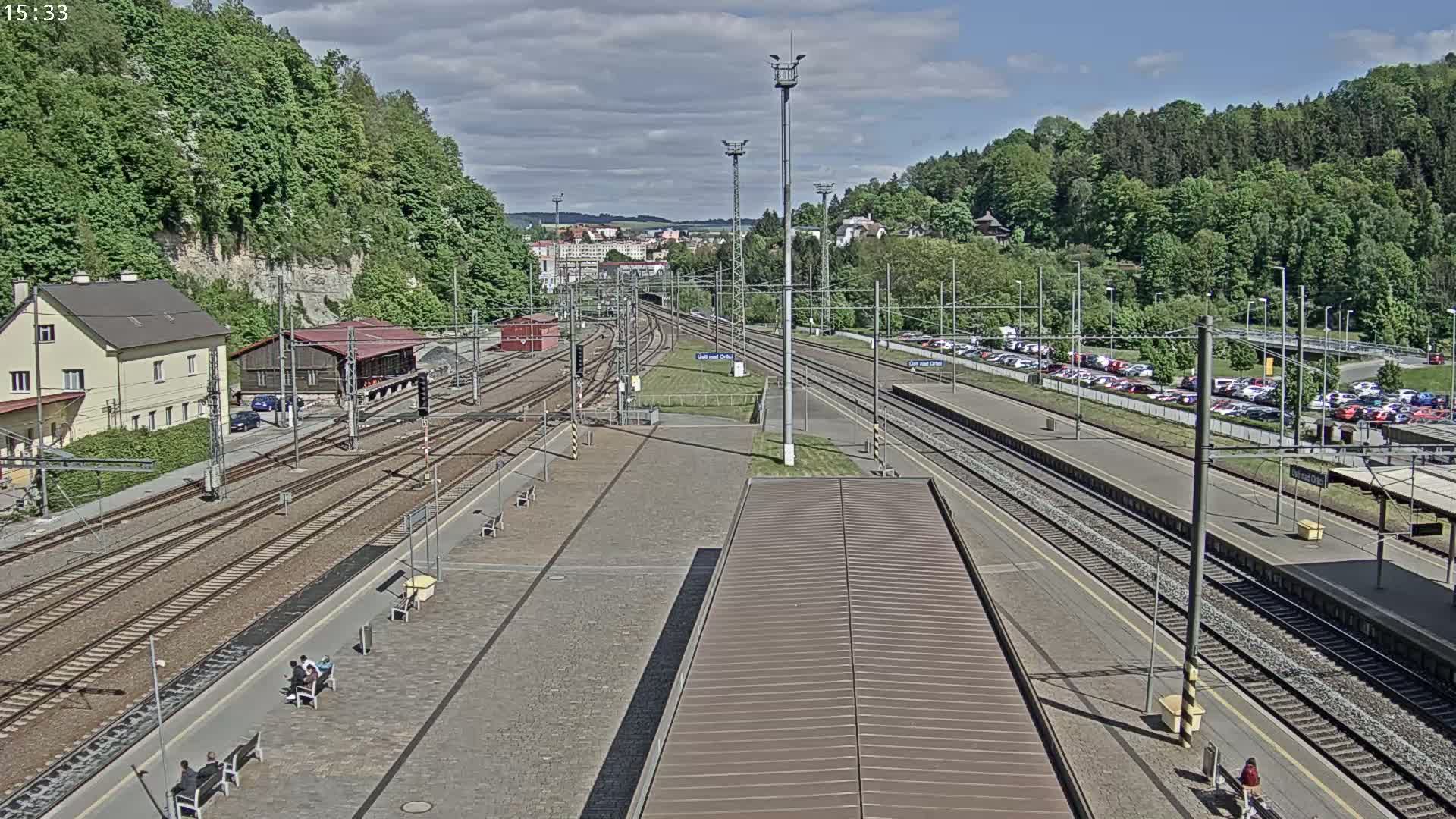 An elevated view of a train station on a partly cloudy day, showing multiple train tracks, platforms with people sitting on benches, a parking lot full of cars, and lush green hills in the background.