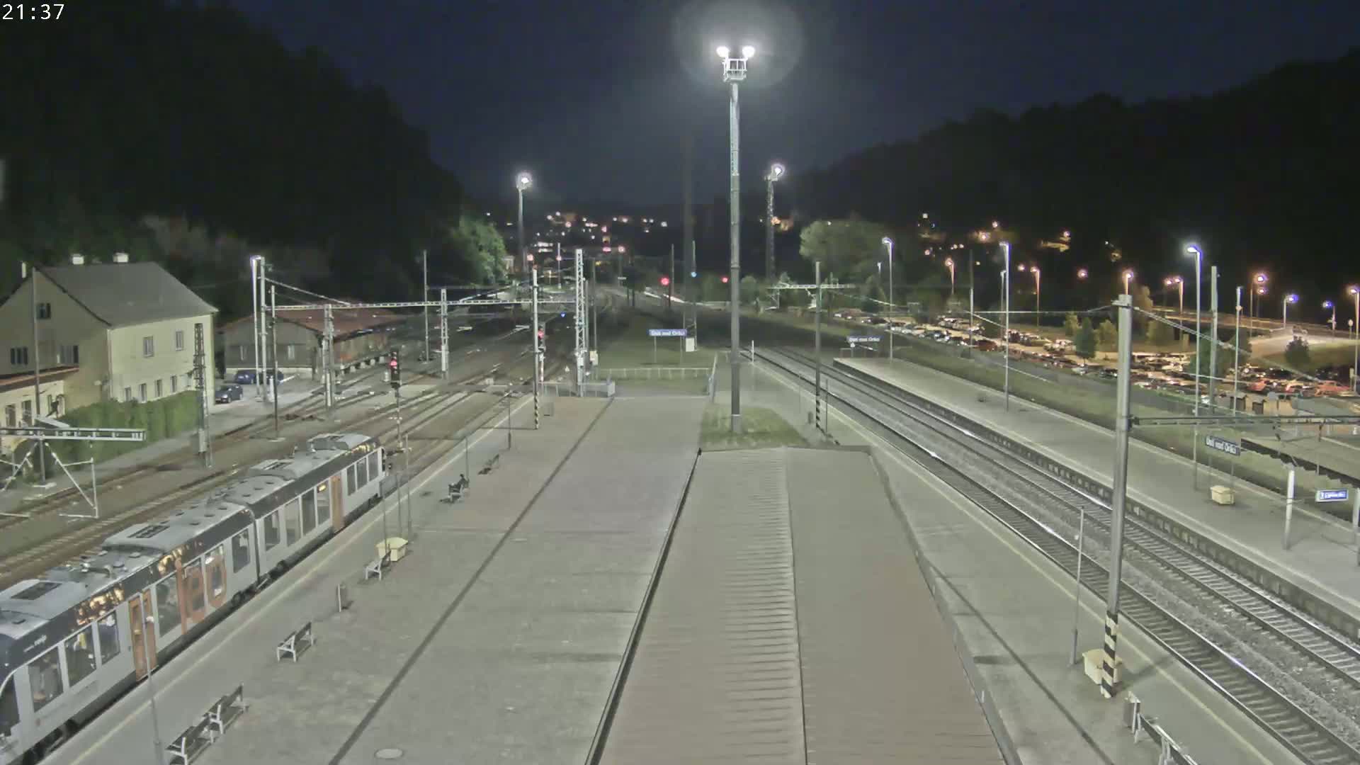 A nighttime view of a train station with a train at the platform, multiple tracks, and a parking lot filled with cars, all surrounded by dark hills.