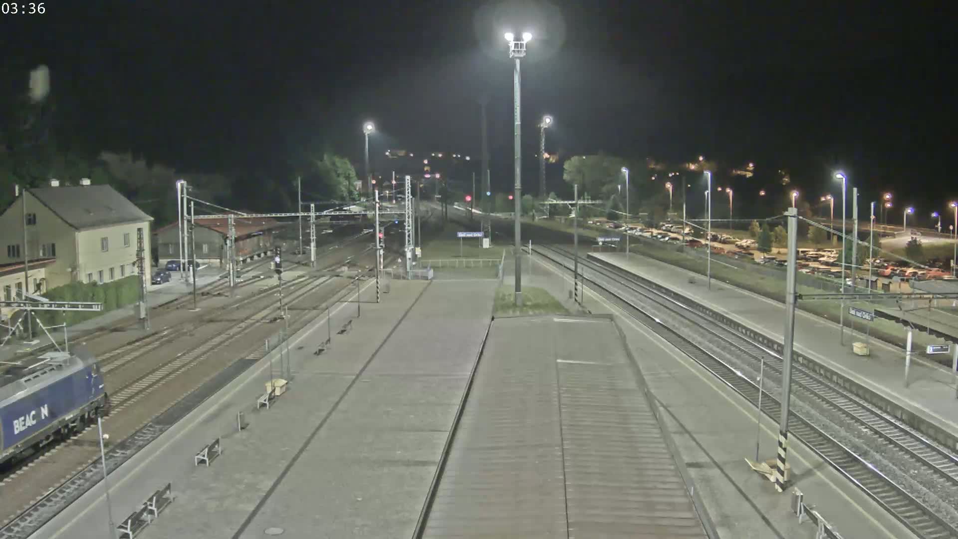 A nighttime view of a train station with a blue train partially visible, multiple train tracks, and a parking lot in the background under clear skies.