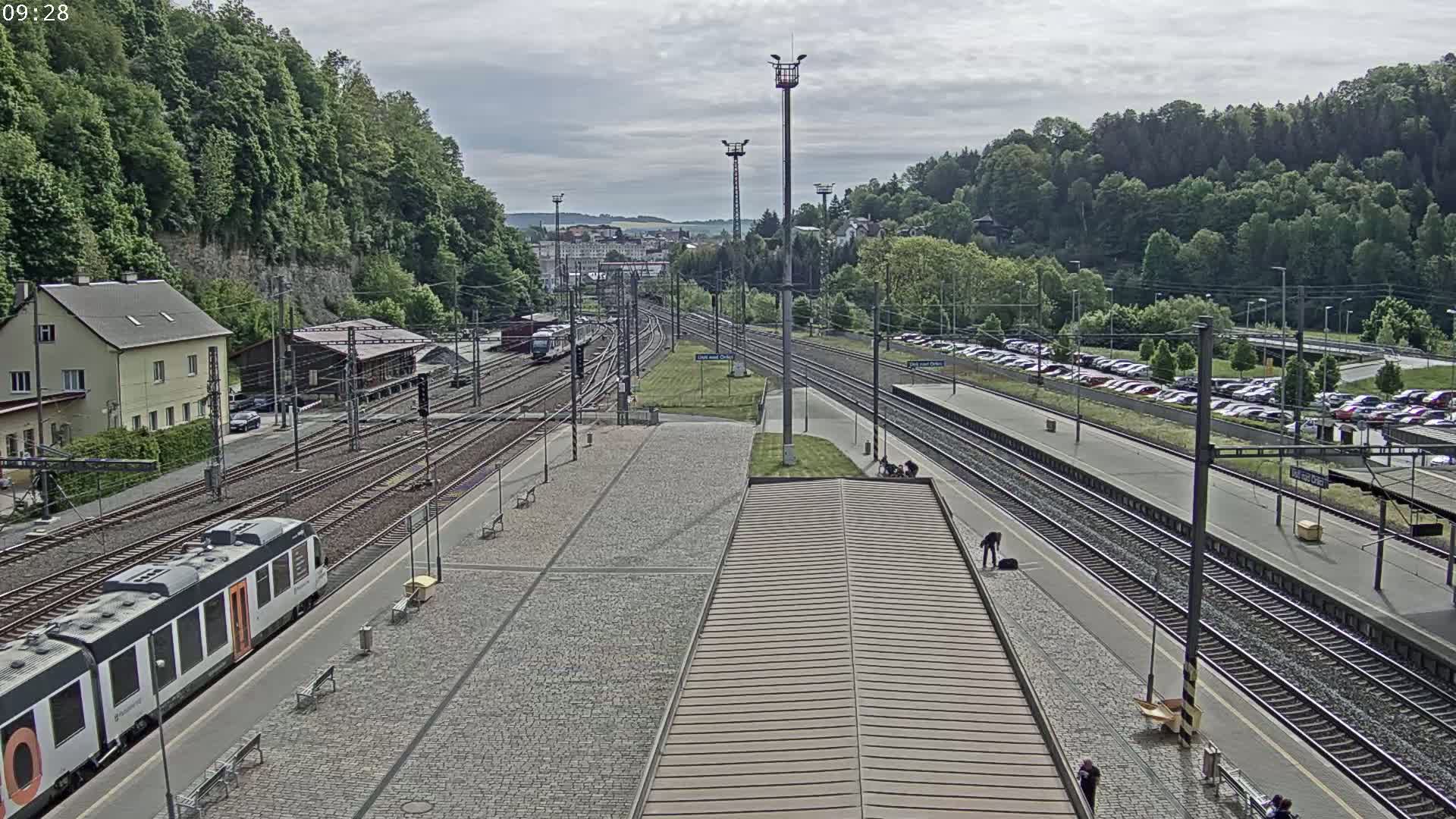 A train station with multiple train tracks, a partially visible train, and a large parking lot is seen under an overcast sky.