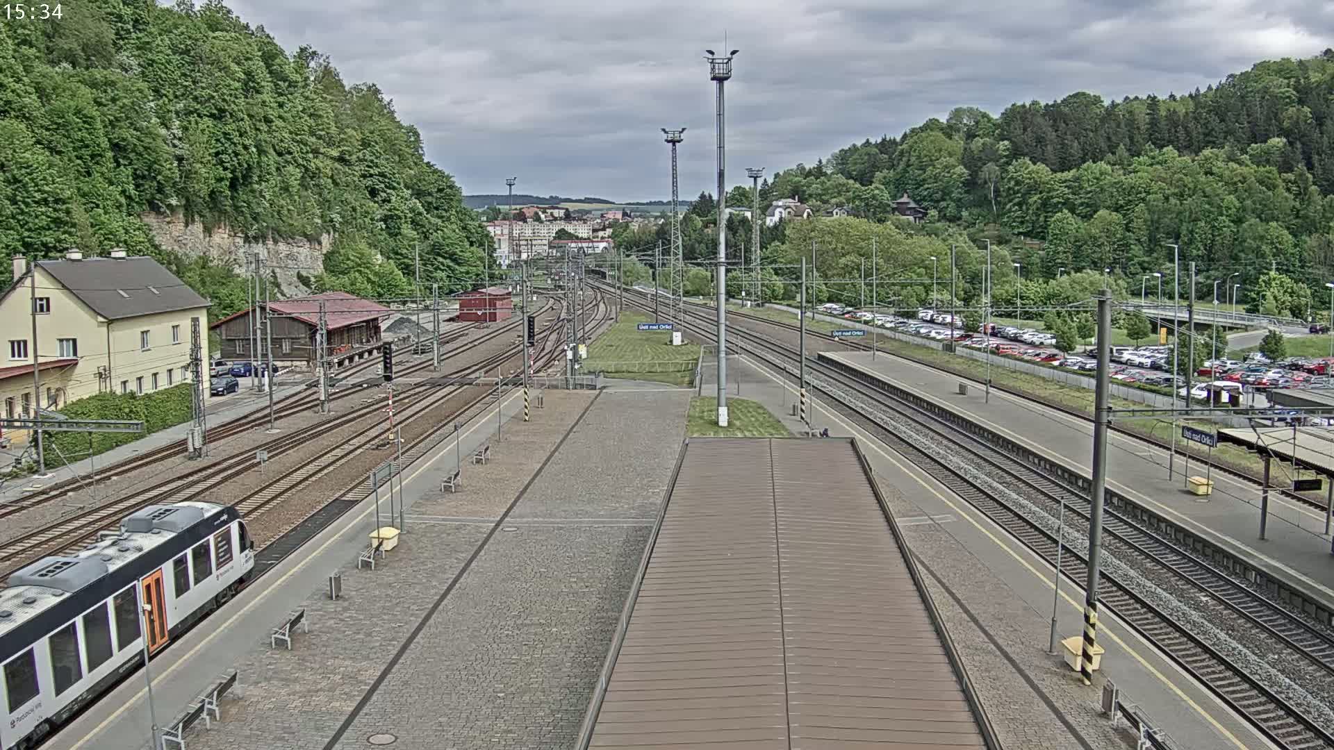 A train station with multiple tracks, a train, and several buildings sits under a cloudy sky, surrounded by lush green hills.