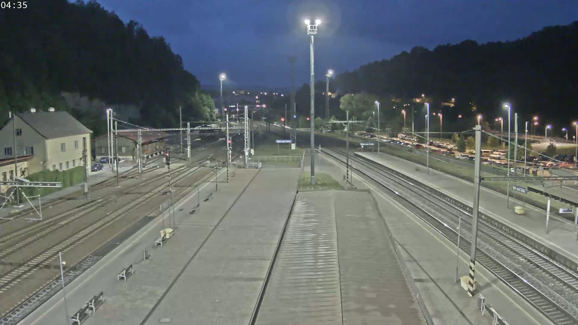 A nighttime view of a train station platform with multiple train tracks, a building, and a parking lot in the background under a dark sky.
