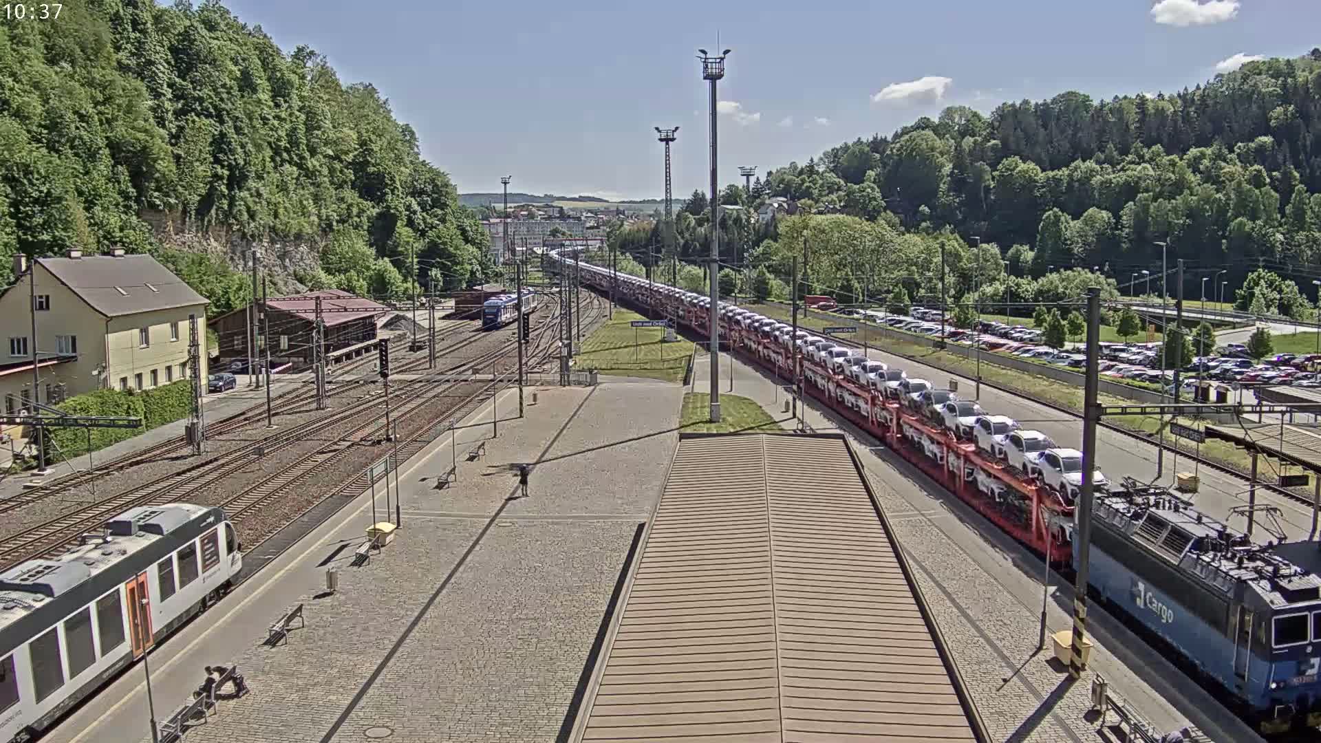 A long train carrying numerous cars sits in a train station on a sunny day, with lush green hills in the background.