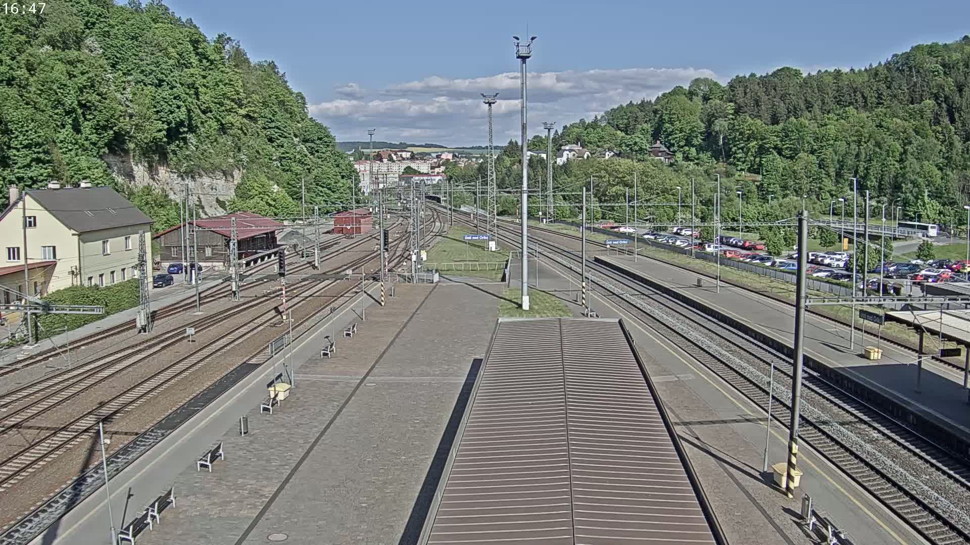 A train station with multiple tracks and a large parking lot is seen on a sunny day, surrounded by lush green hills.