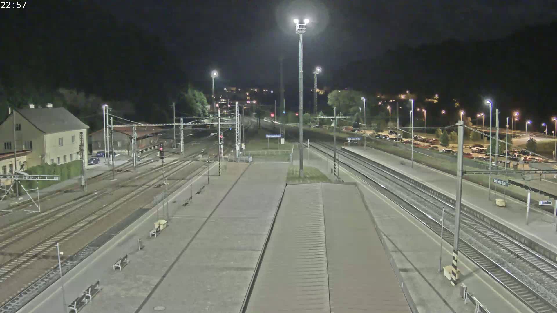A nighttime view of a train station with multiple train tracks, platforms, and a parking lot, illuminated by artificial lights.