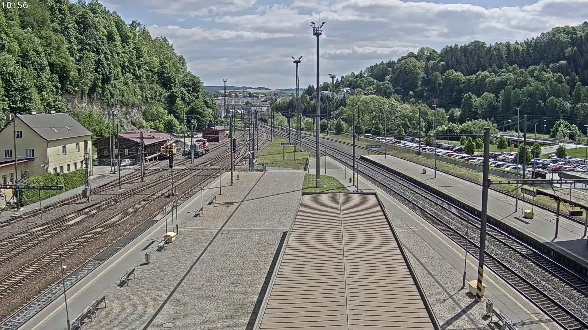 A train station with multiple train tracks, a building, and a large parking lot is visible under a partly cloudy sky.