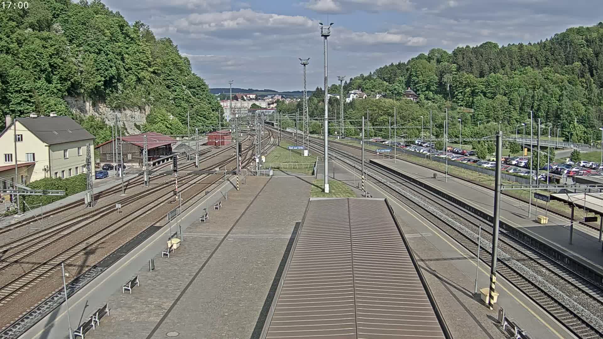 A high-angle view of a train station on a partly cloudy day, showing multiple train tracks, a parking lot filled with cars, and a building nestled against a hillside covered in lush green trees.