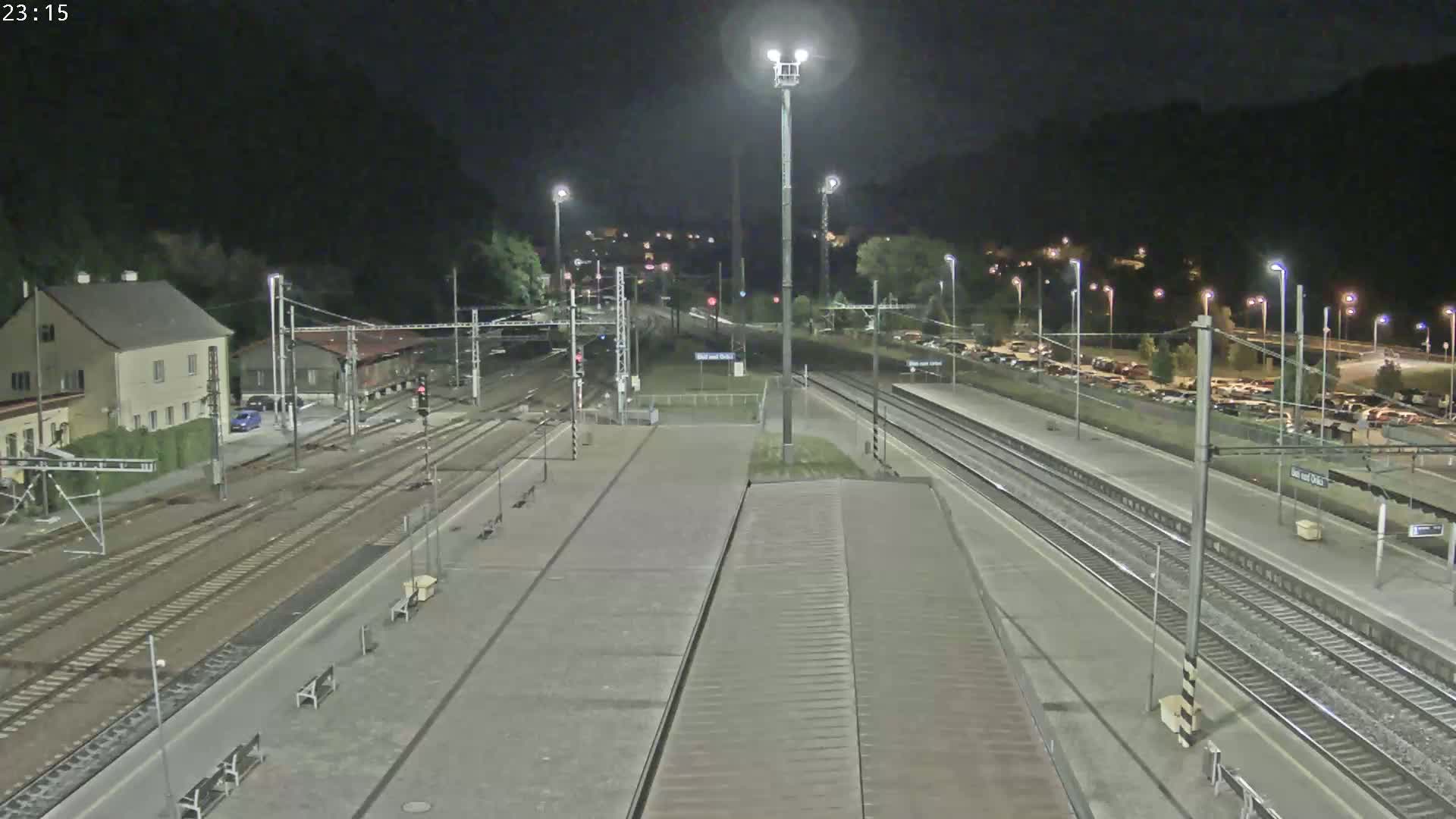 A nighttime view of a train station with multiple train tracks, a platform, and a parking lot, illuminated by artificial lights.