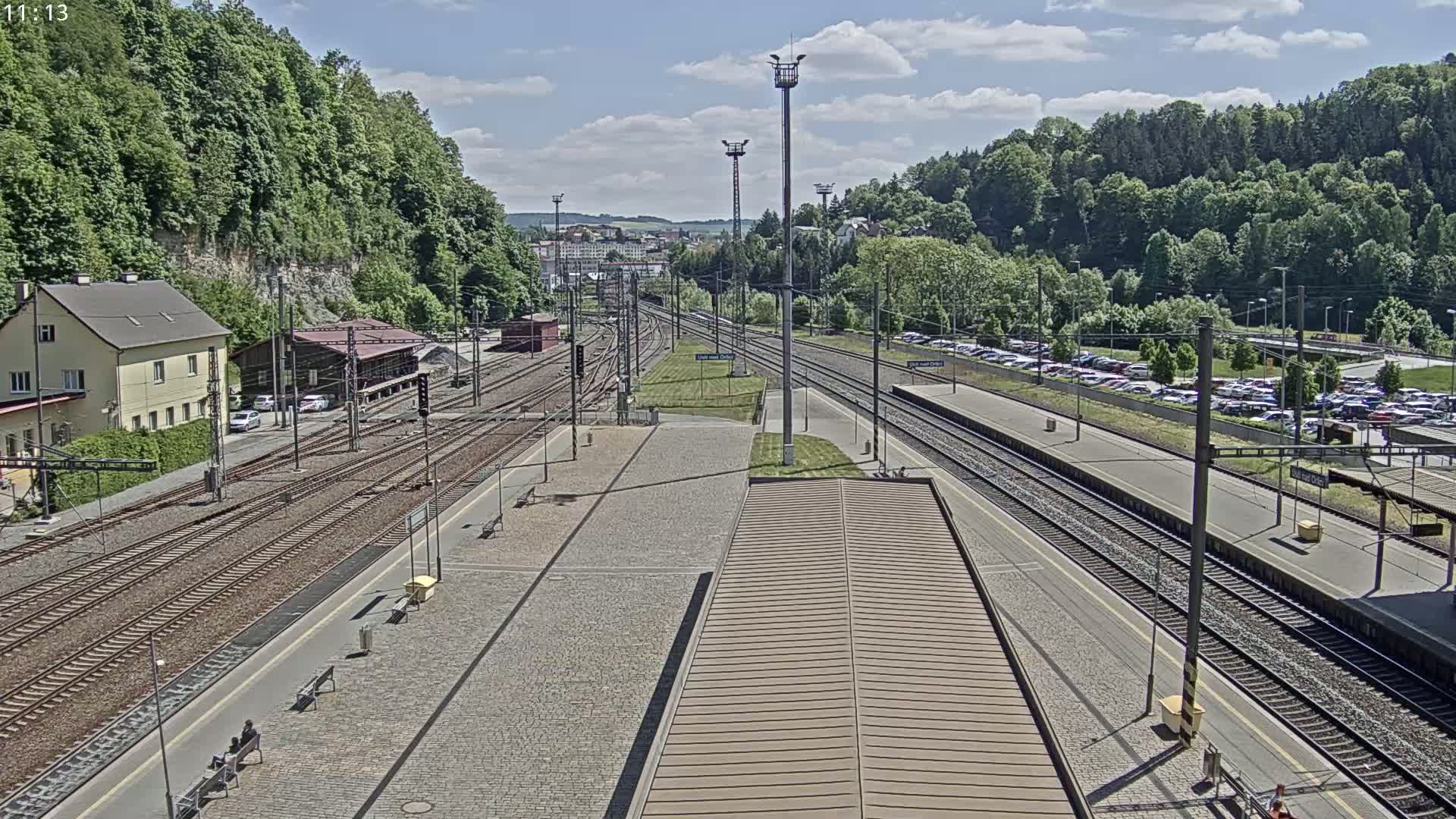 A sunny day at a train station with multiple tracks, a large parking lot, and lush green hills in the background.