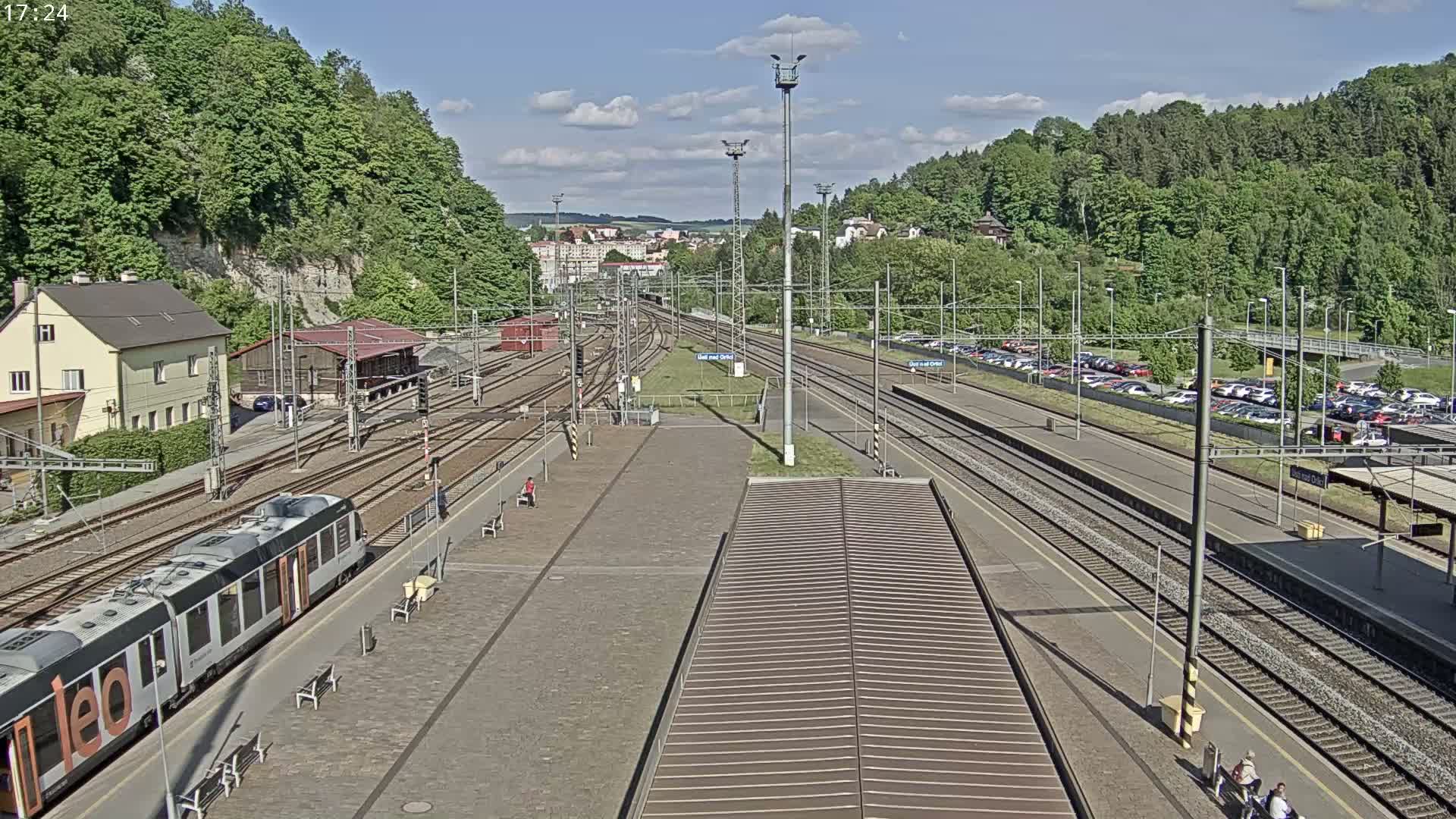 A train station on a sunny day with multiple train tracks, a passenger train, and a large parking lot is surrounded by green hills.