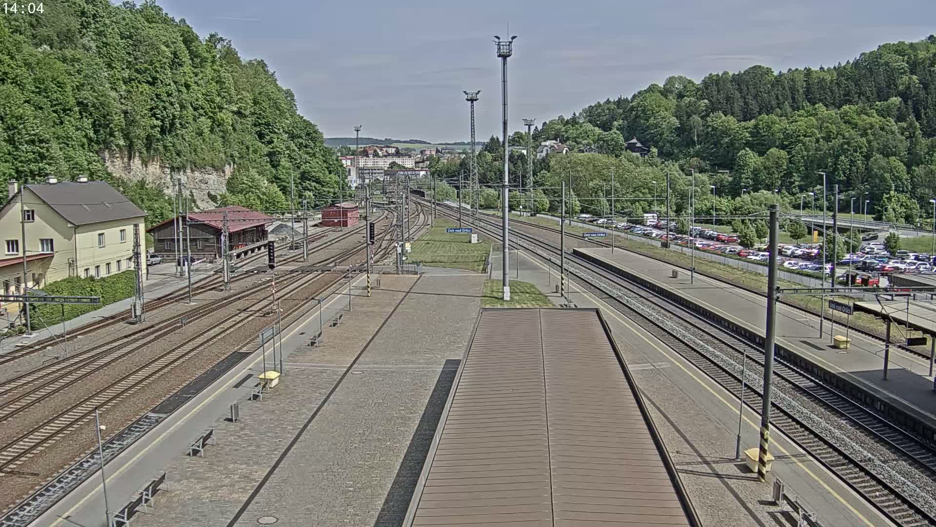 A train station with multiple tracks, a large parking lot, and lush green hills in the background on a sunny day.