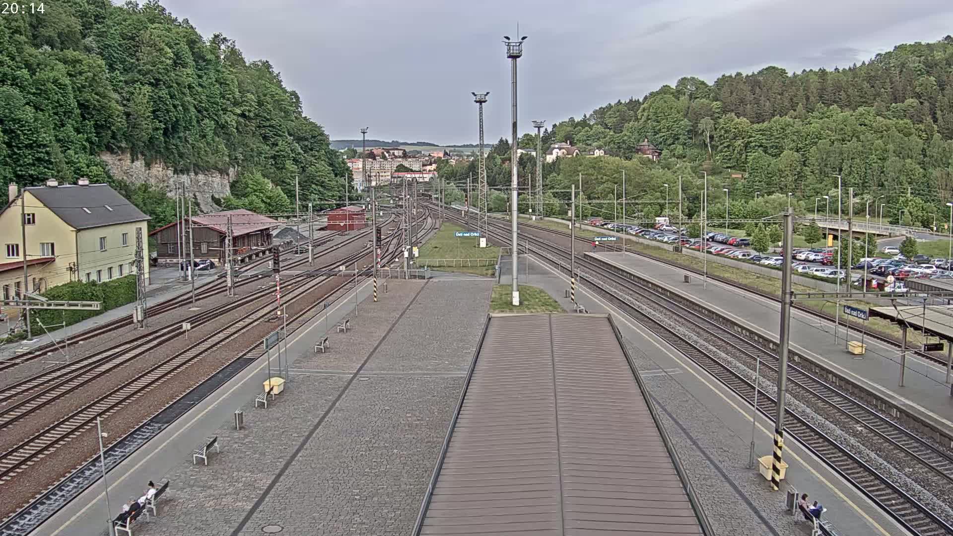 A train station with multiple tracks, a paved platform, and a small building is situated on a hillside surrounded by lush green trees under an overcast sky.