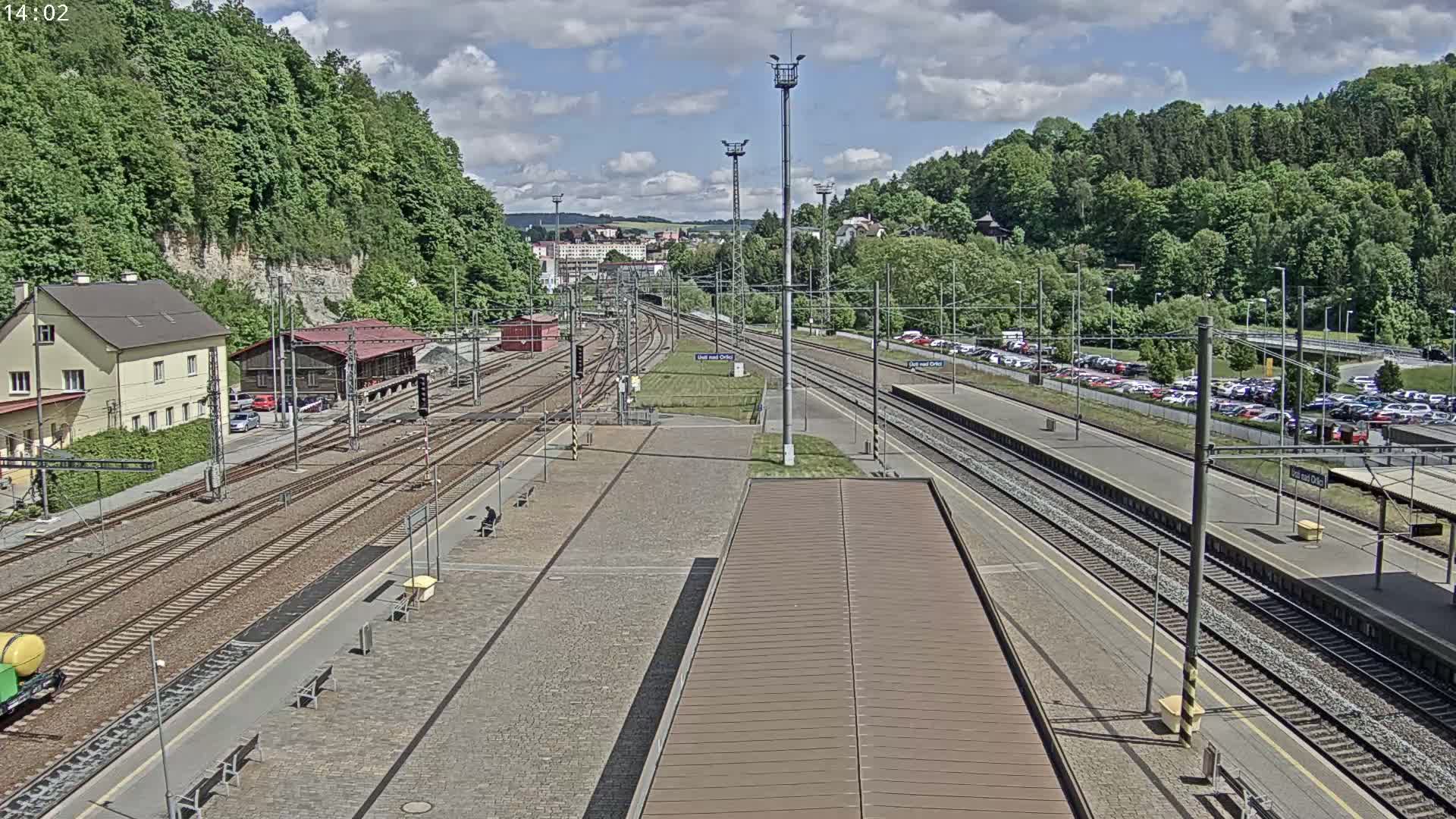 A train station with multiple tracks, surrounded by lush green hills under a partly cloudy sky.