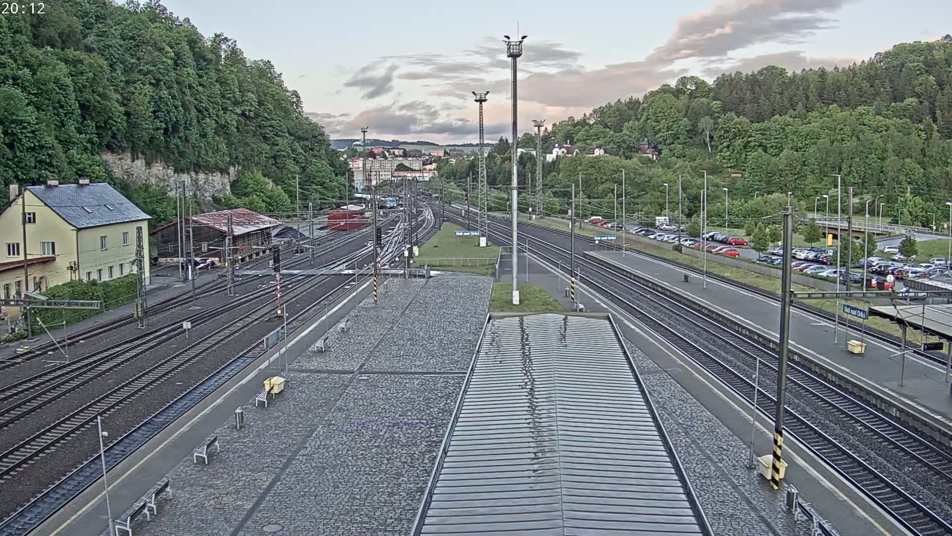 A high-angle view of a train station on an overcast day, showing multiple train tracks, platforms, a parking lot, and buildings surrounded by lush green hills.