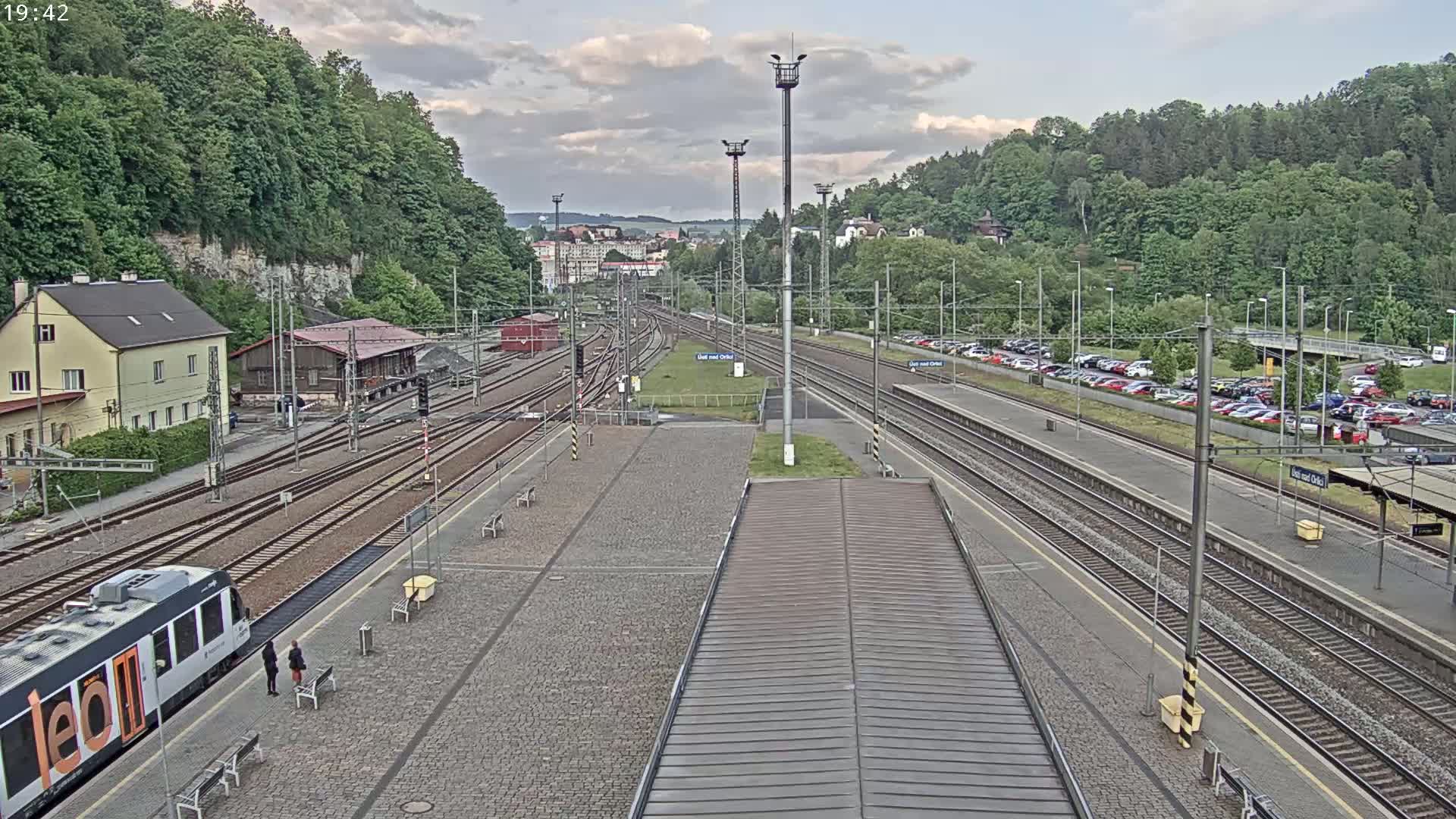 A train station with multiple train tracks, a passenger train, a building, and a large parking lot is visible under partly cloudy skies.