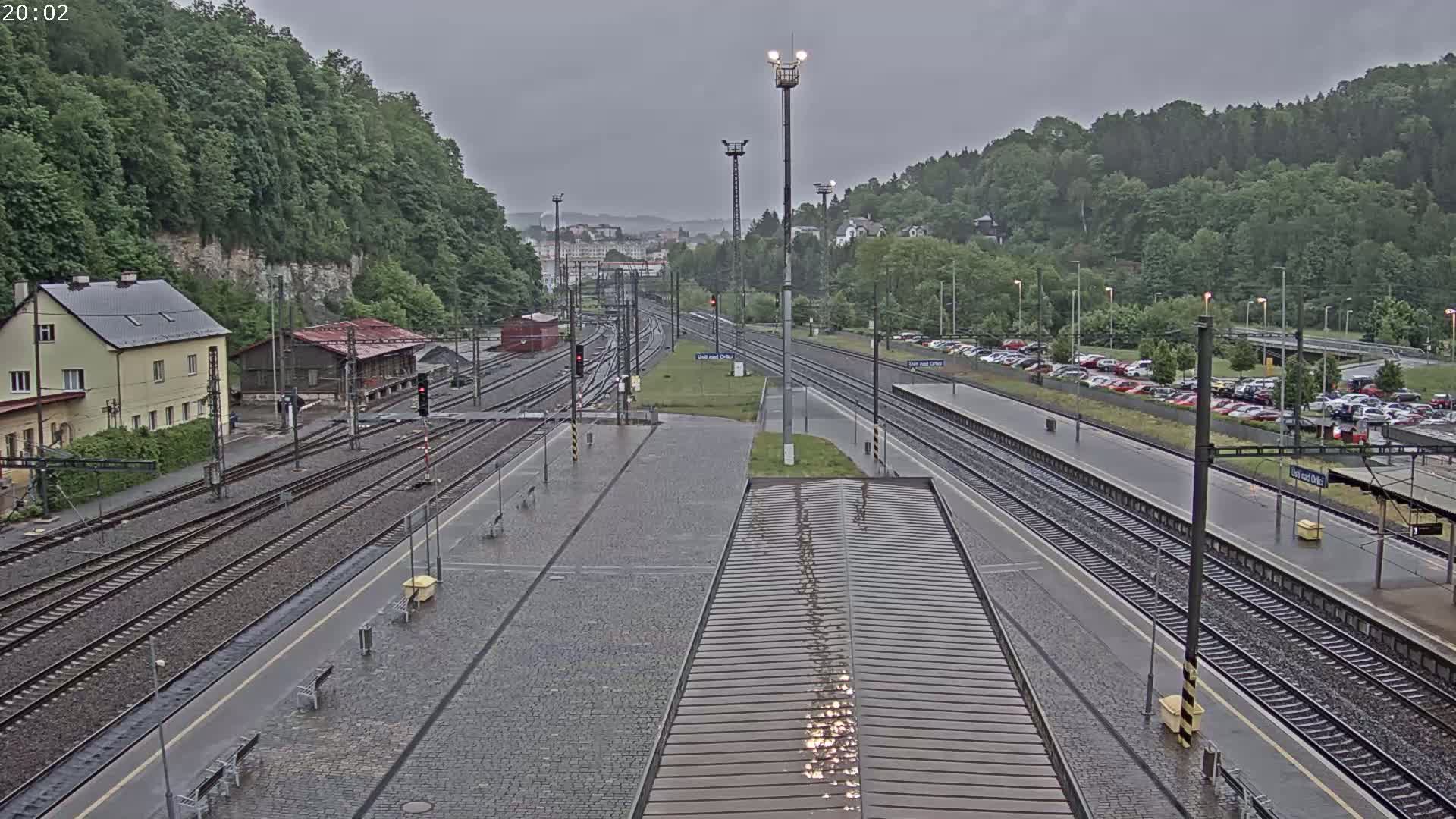 A rain-slicked train station with multiple tracks, surrounded by lush green hills under an overcast sky.