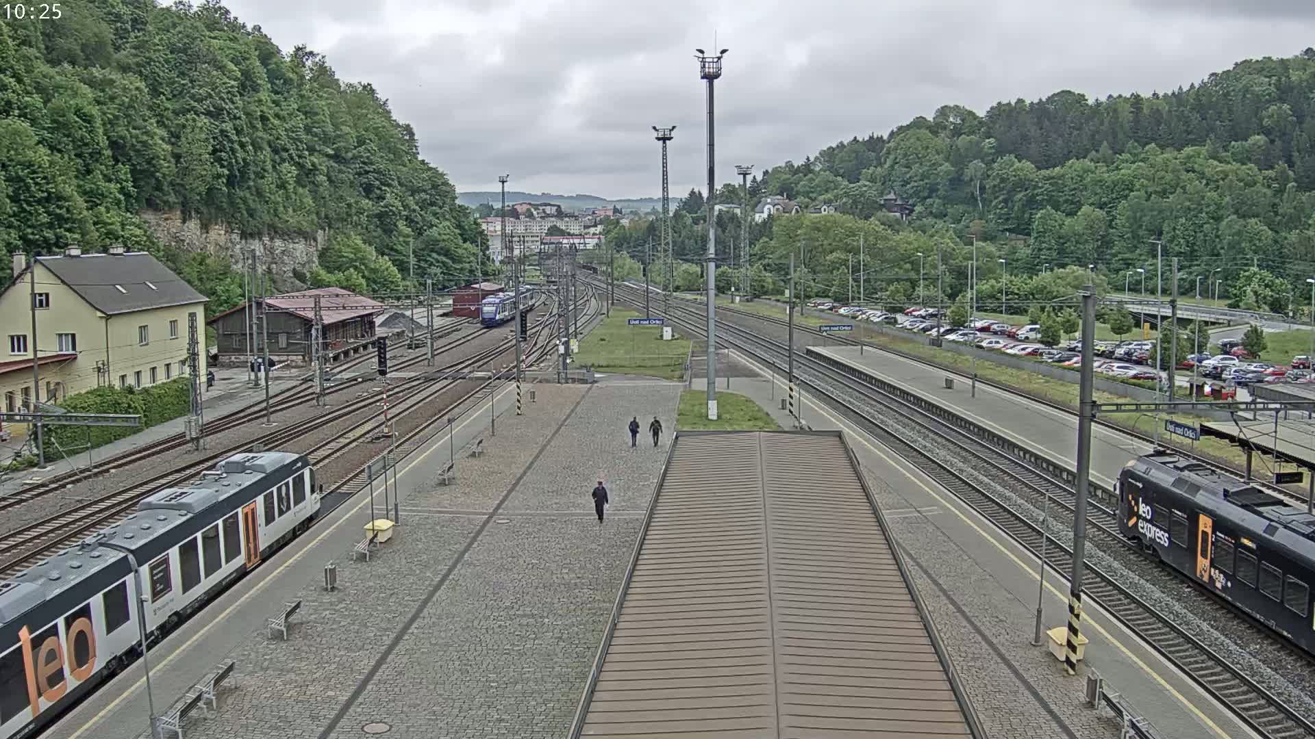 A train station on an overcast day, with multiple train tracks, several trains, and a large parking lot visible.