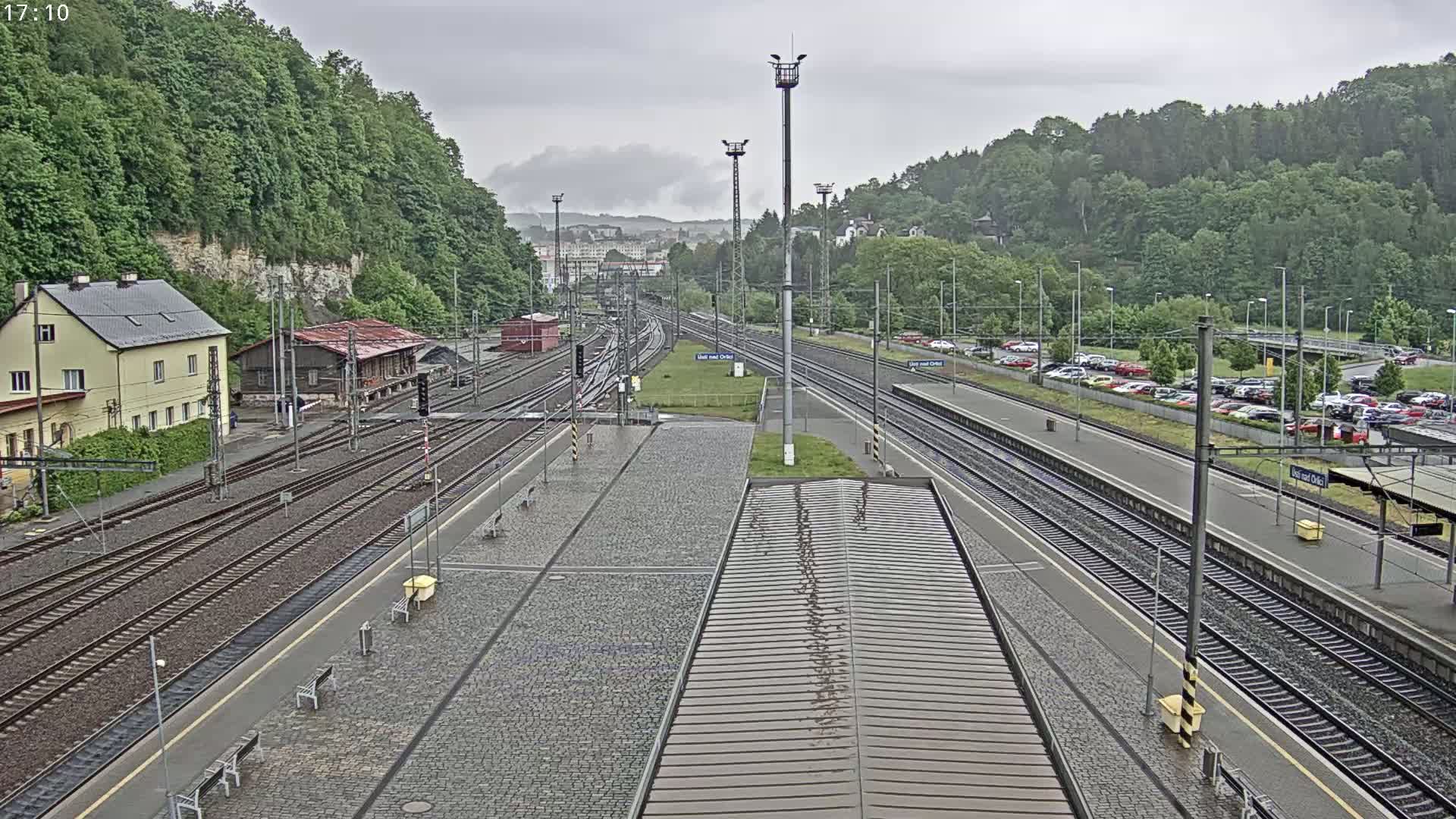 A wet train station with multiple tracks and a large parking lot is surrounded by lush green hills under an overcast sky.