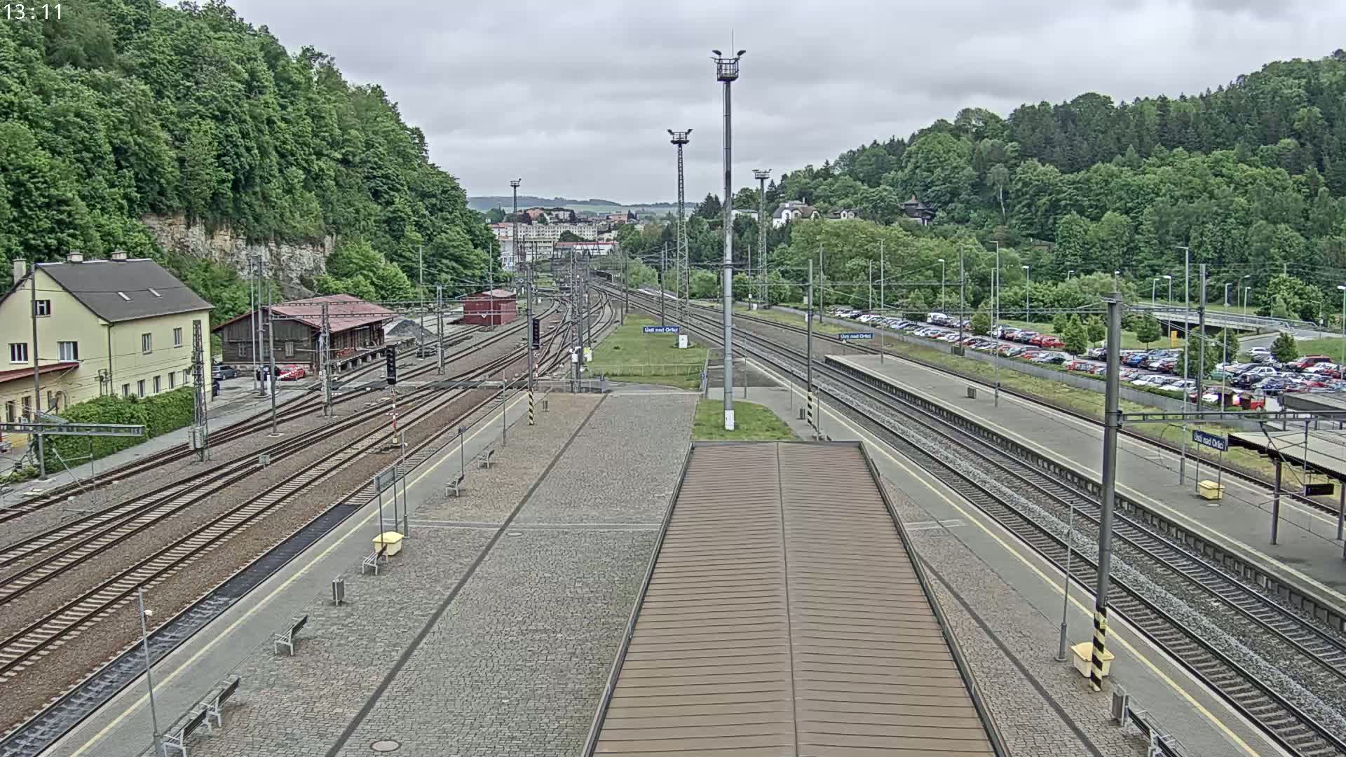 A train station with multiple tracks, a large parking lot, and lush green hills in the background under an overcast sky.