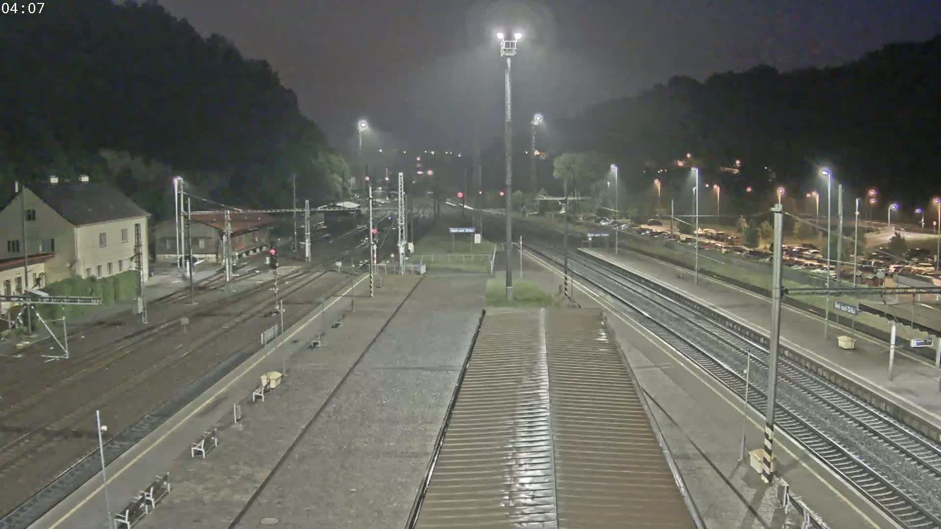 A nighttime view of a rain-slicked train station with multiple tracks and a building in the background.