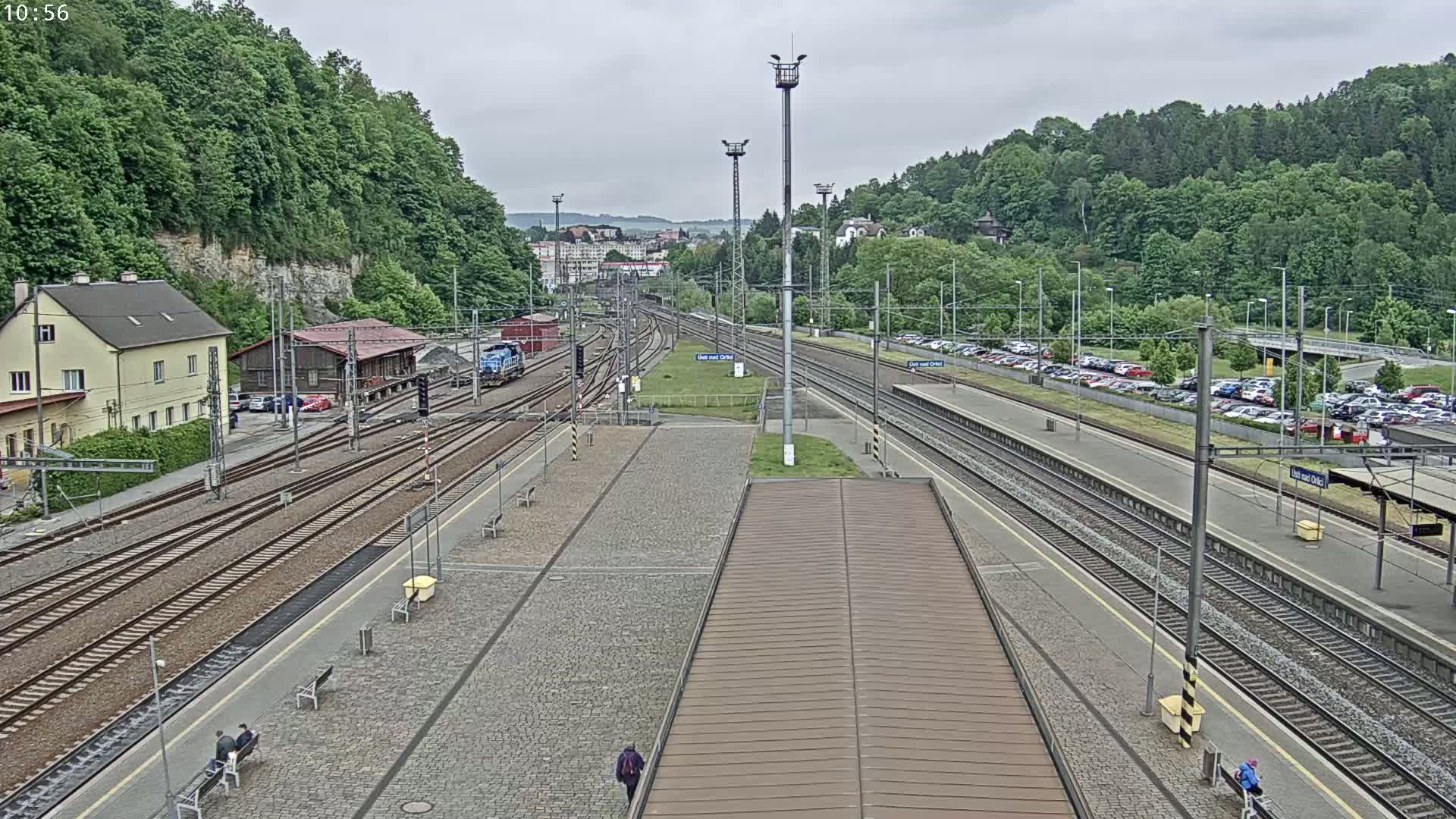 A train station with multiple train tracks, a large parking lot, and surrounding green hills under an overcast sky.