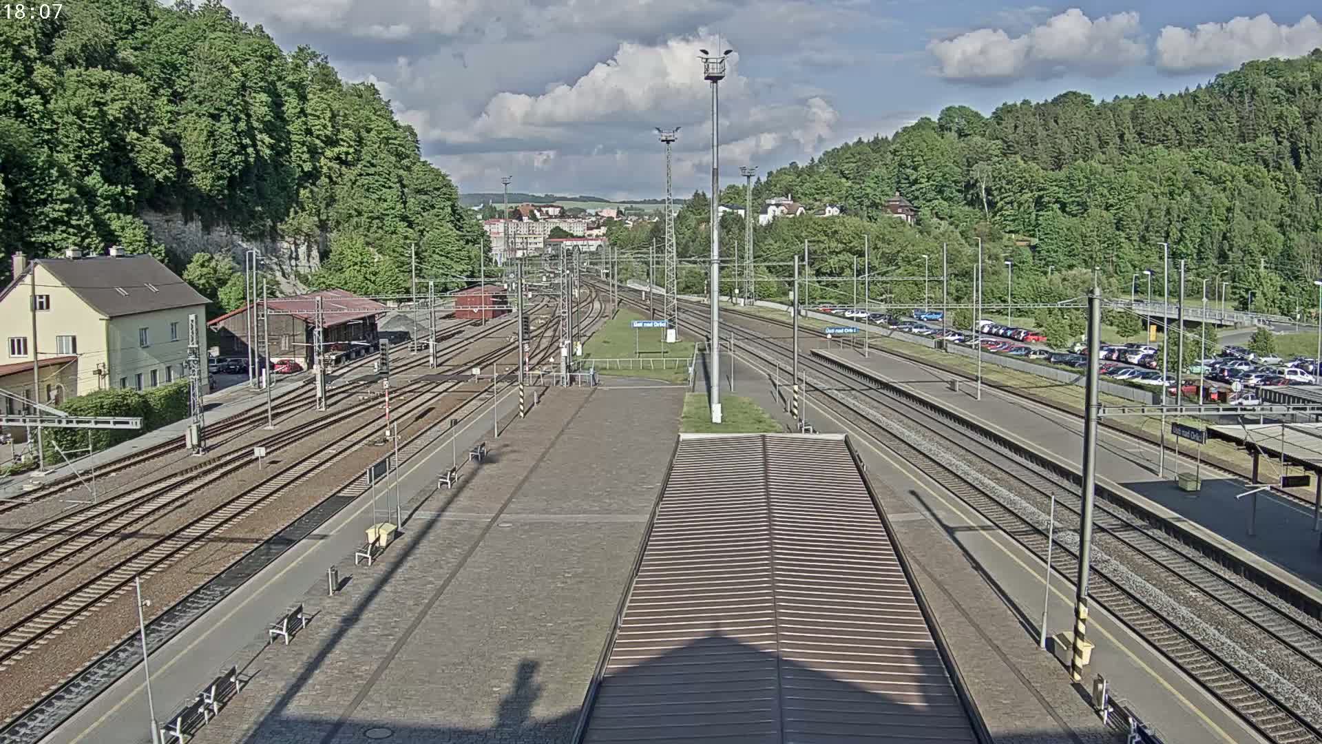A high-angle view of a train station on a partly cloudy day, showing numerous train tracks, platforms, a building, and a parking lot filled with cars, all surrounded by lush green hillsides.