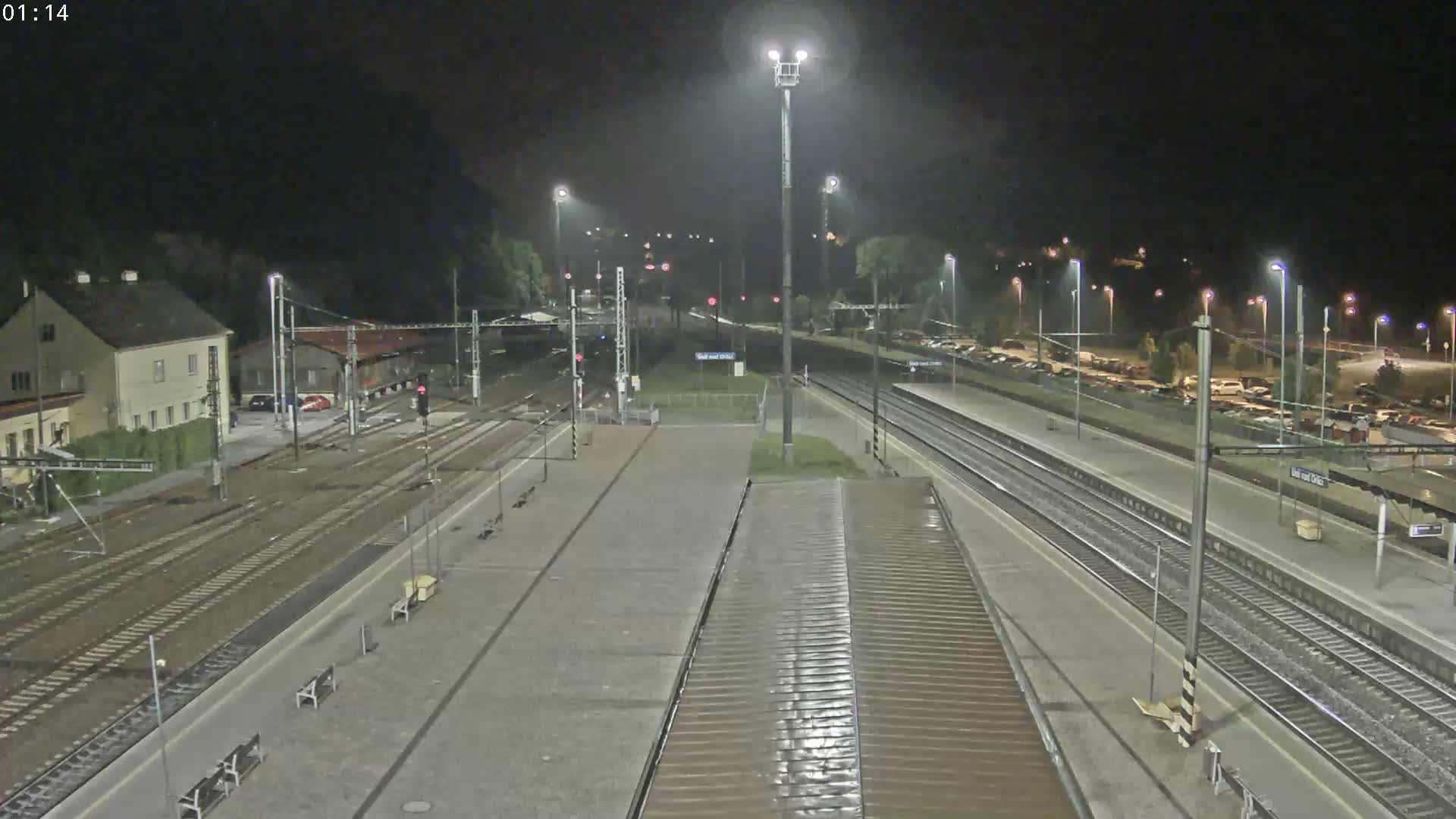 A nighttime view of a train station with multiple tracks, platforms, and a parking lot, under dark skies.