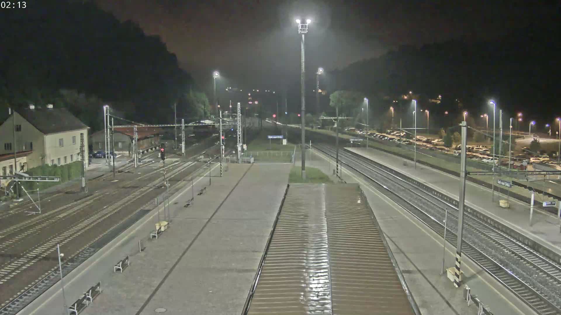 A nighttime view of a train station with multiple tracks, a platform, and a parking lot, illuminated by artificial lights under a dark, possibly overcast sky.