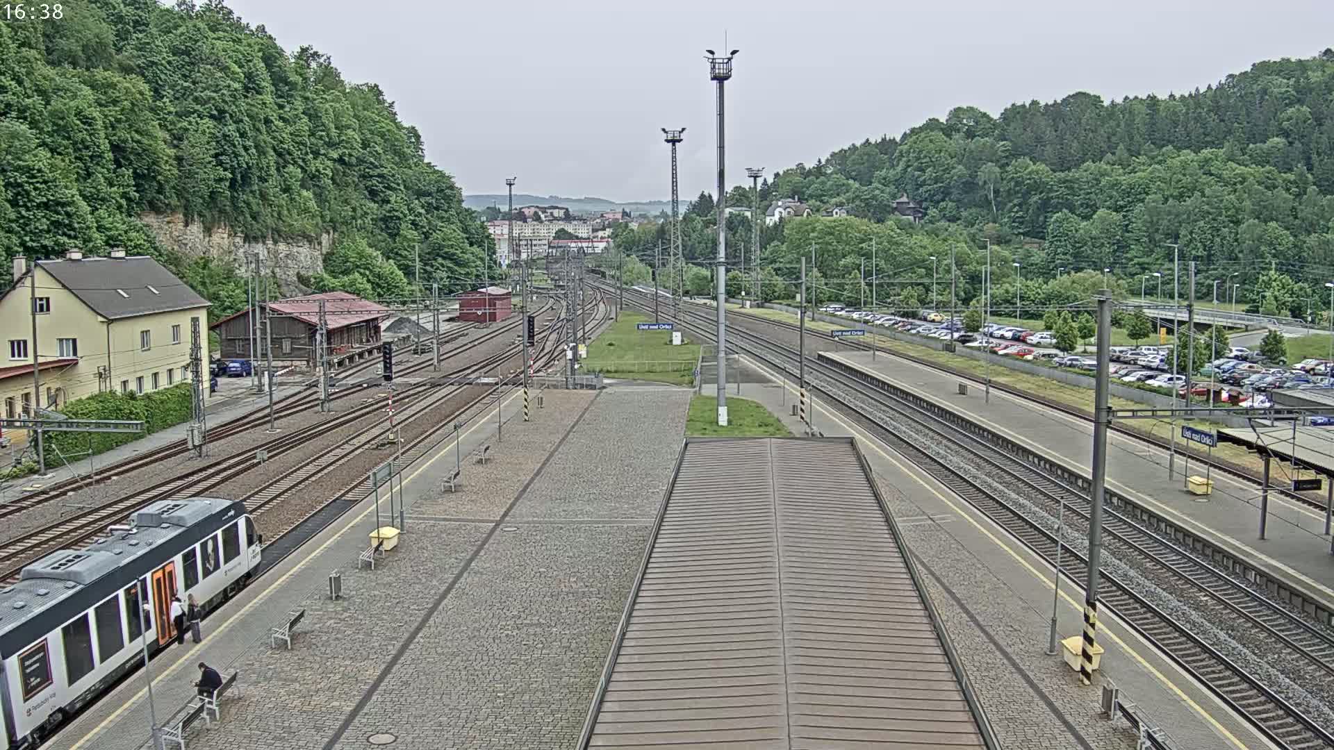 An overcast day at a train station with multiple tracks, a passenger train at the platform, a large parking lot, and lush green hills in the background.