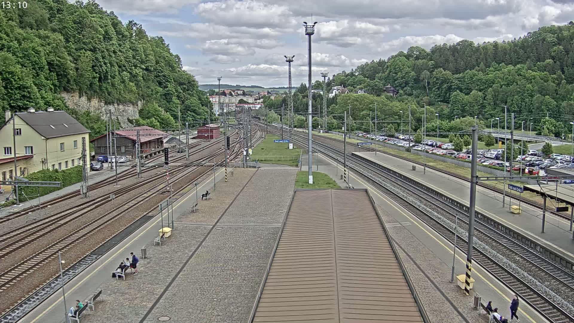 A train station with multiple tracks, a large parking lot, and several people sitting on benches under a partly cloudy sky.