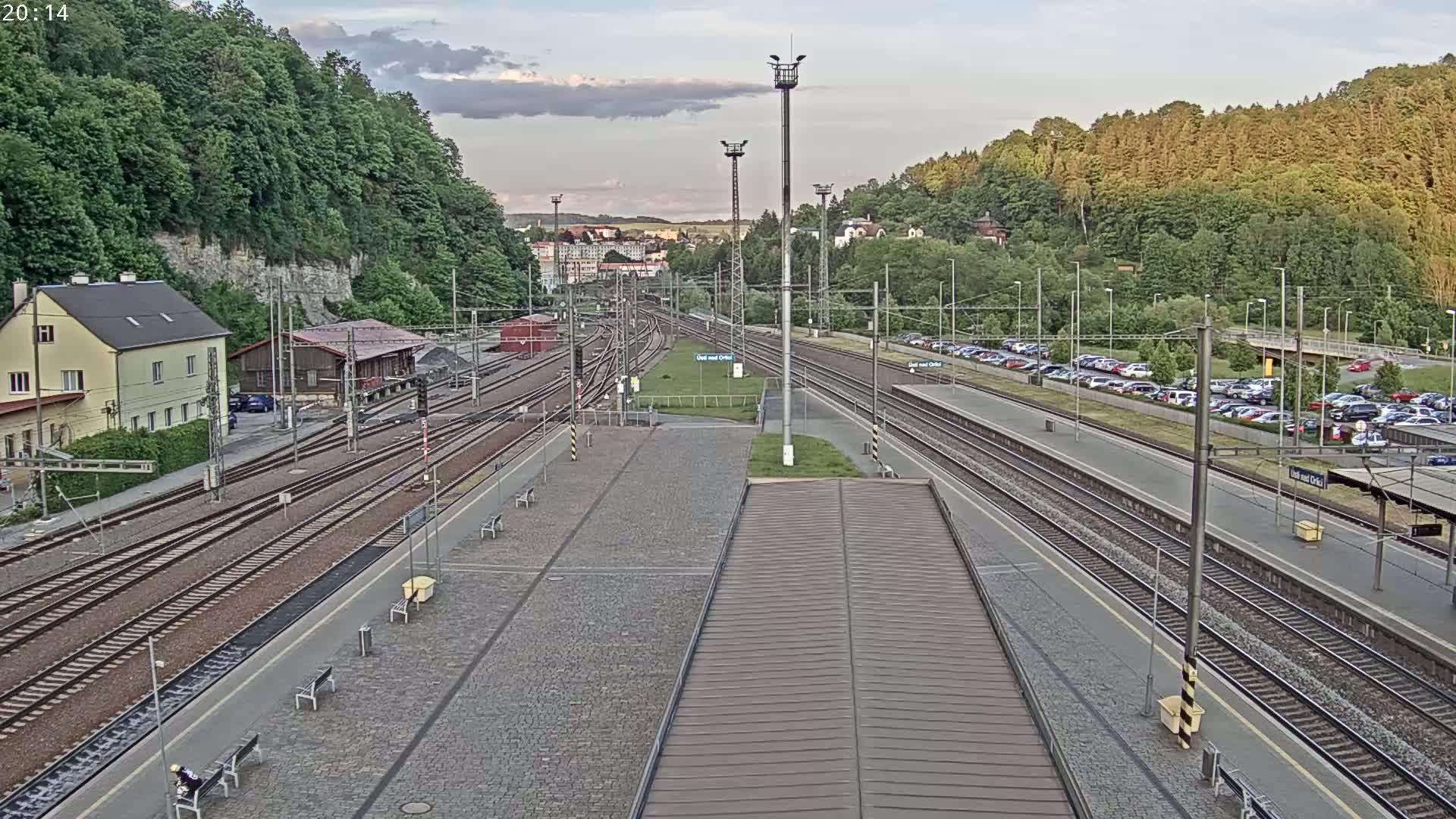 A train station with multiple tracks and a large parking lot is seen under a partly cloudy evening sky.