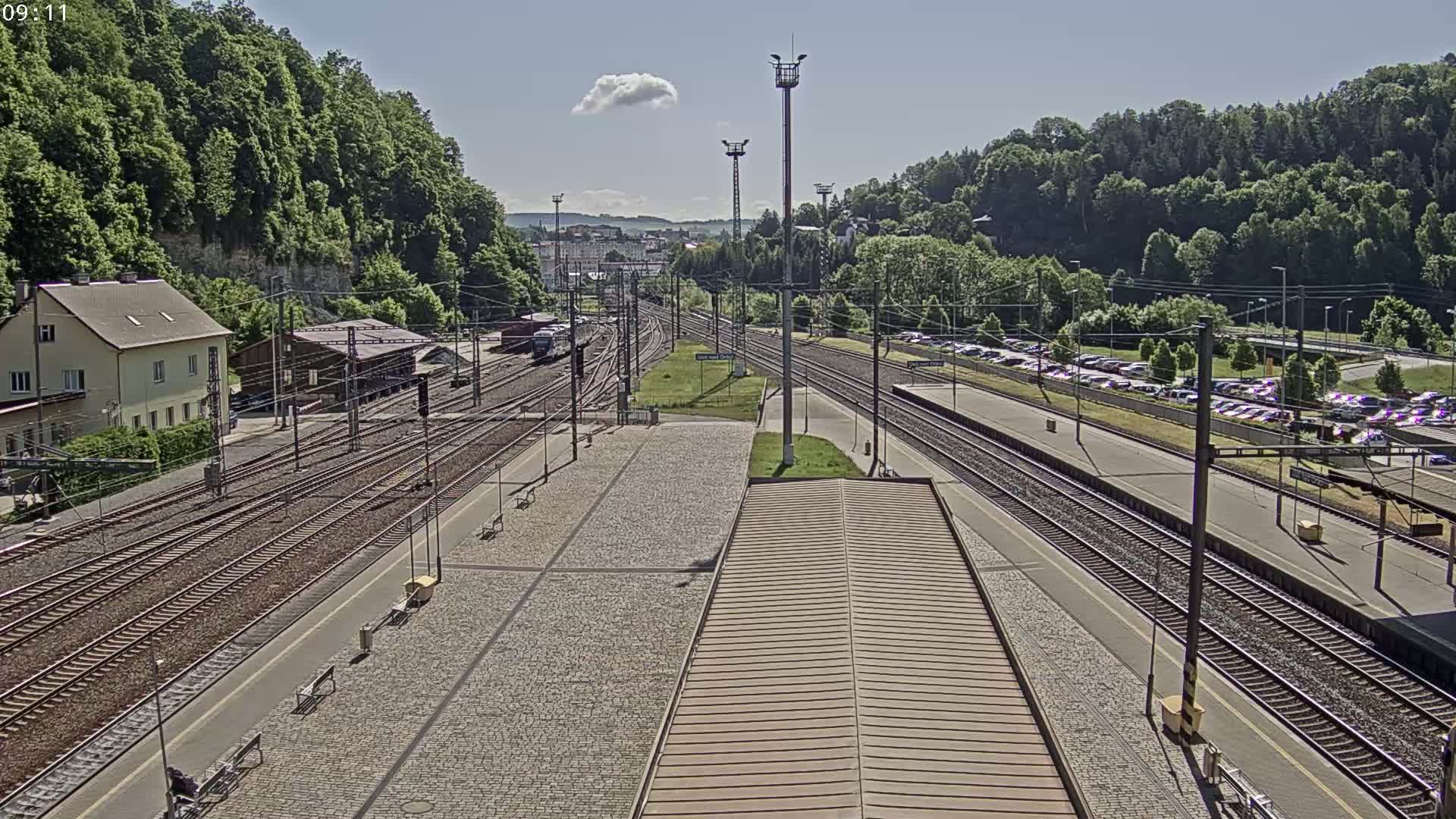A high-angle view of a train station on a sunny day, showing multiple train tracks, a platform with benches, a building, and a large parking lot filled with cars.