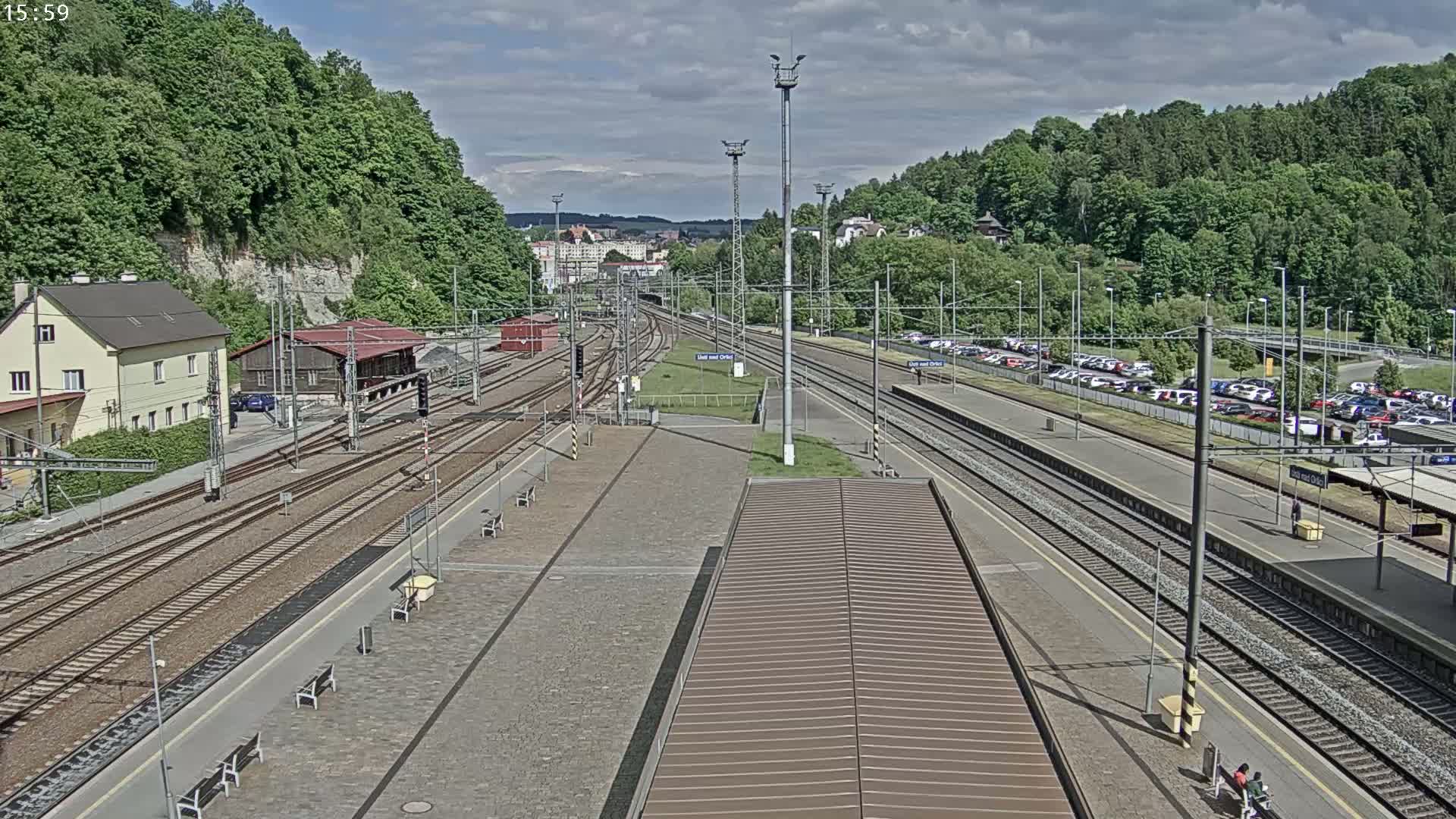A train station with multiple train tracks and a large parking lot is shown under partly cloudy skies.