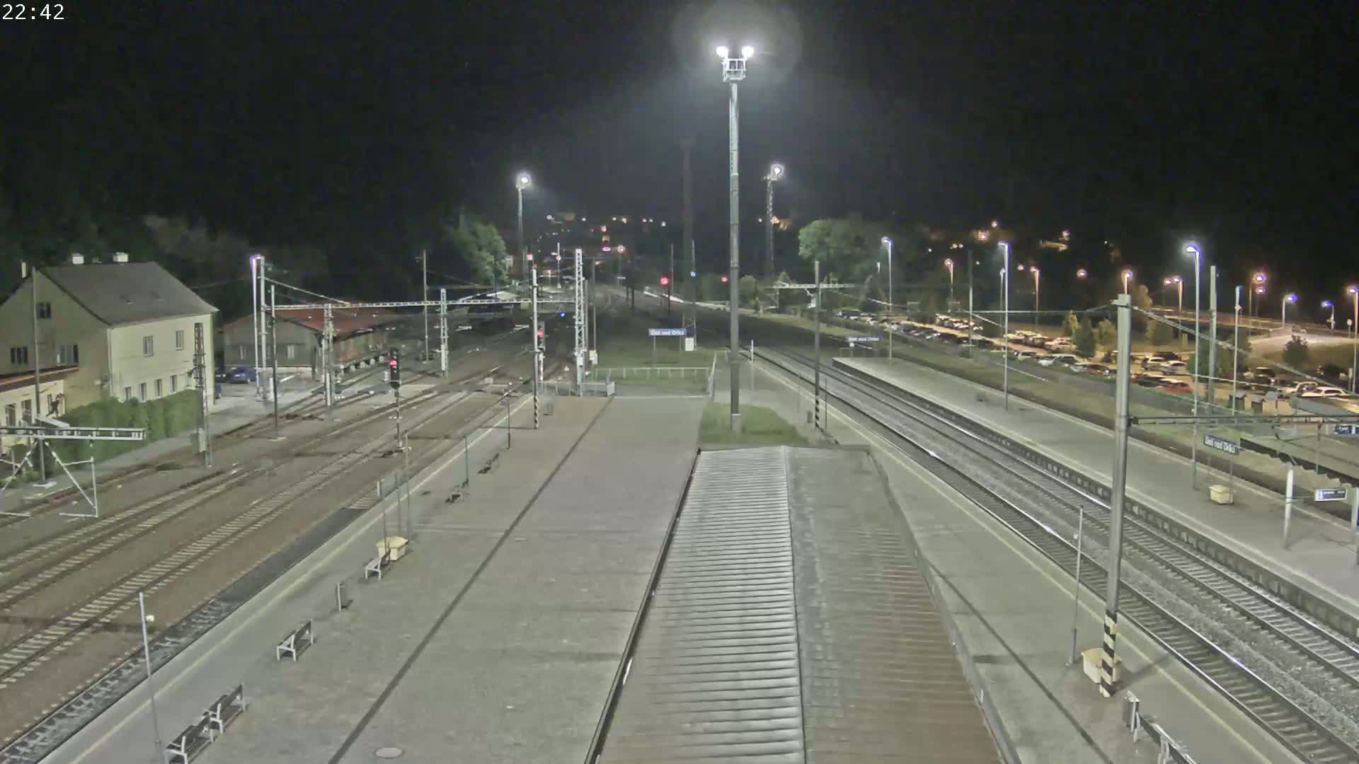 A nighttime view of a train station with multiple tracks, platforms, and a parking lot, illuminated by artificial lights.