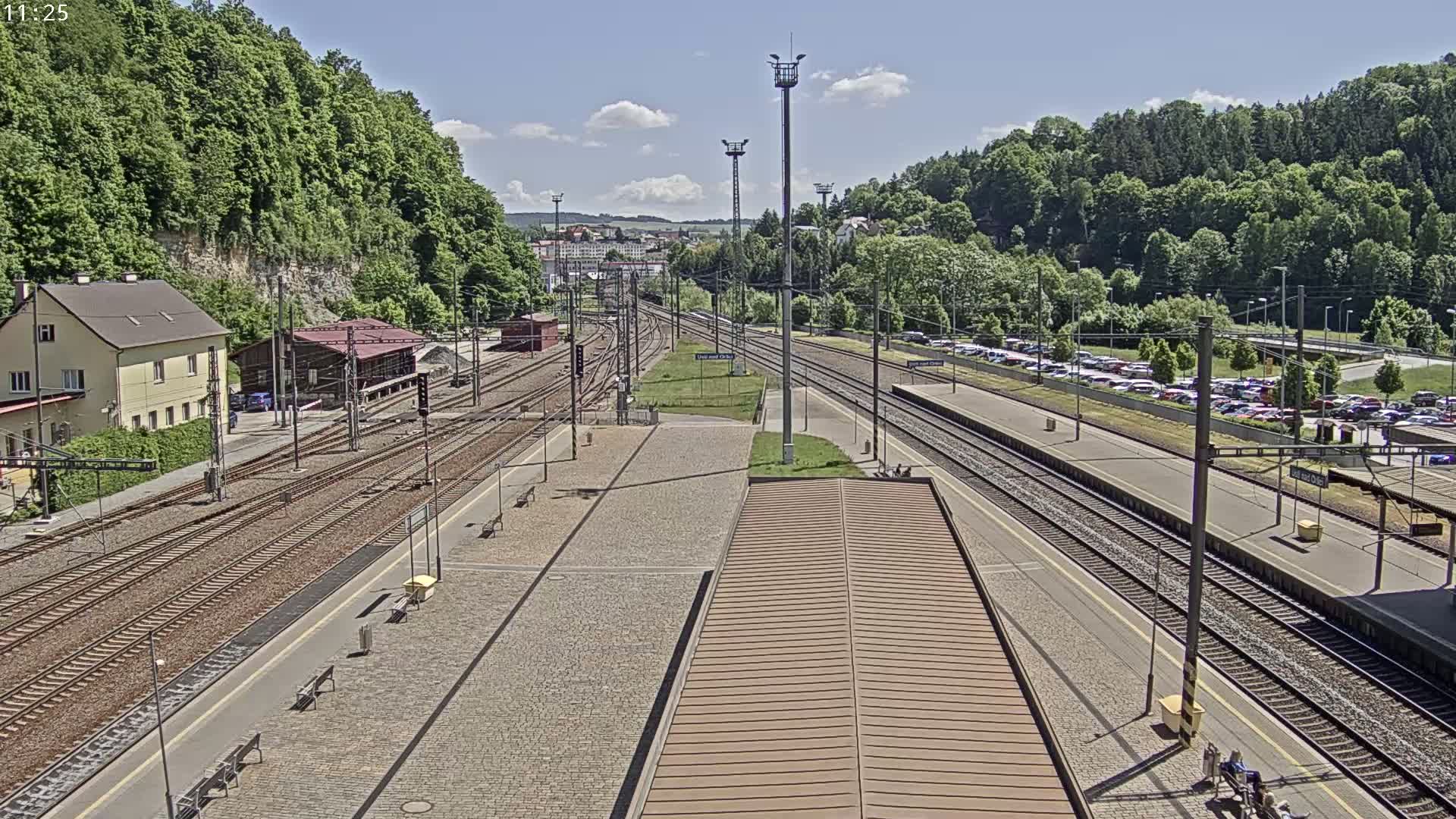 A train station with multiple tracks, a building, and a large parking lot is situated between lush green hills under a mostly sunny sky.