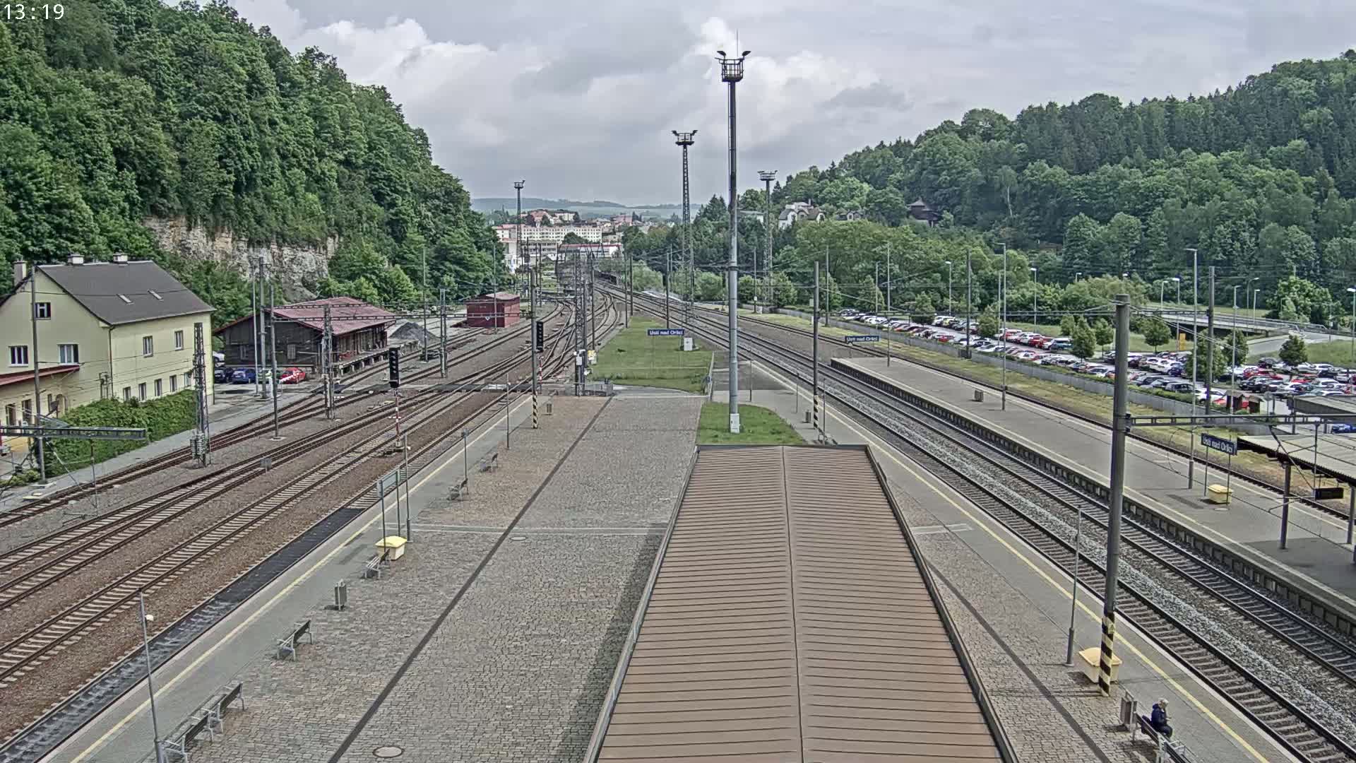 A high-angle view of a train station on an overcast day, showing multiple train tracks, a platform with benches, and a large parking lot filled with cars.