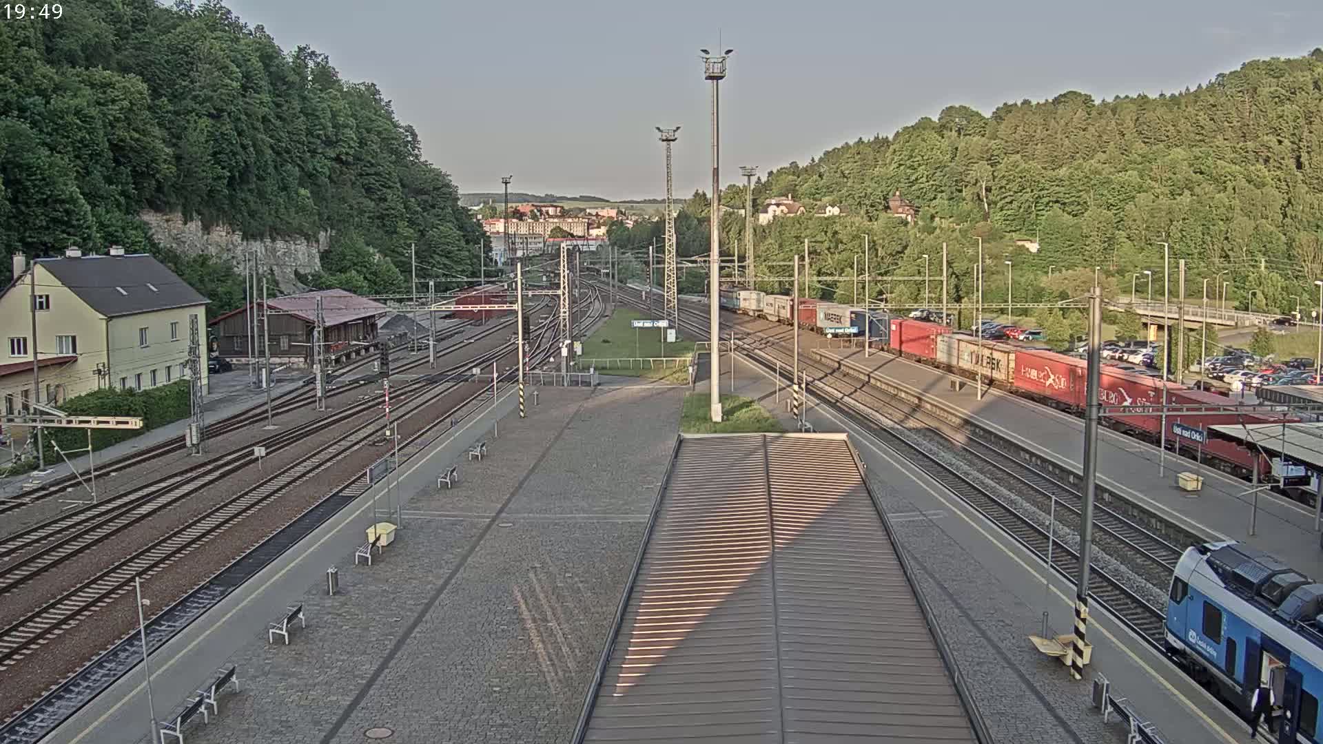 A high-angle view of a train station with multiple train tracks, a few trains, and surrounding green hills under a partly cloudy sky.