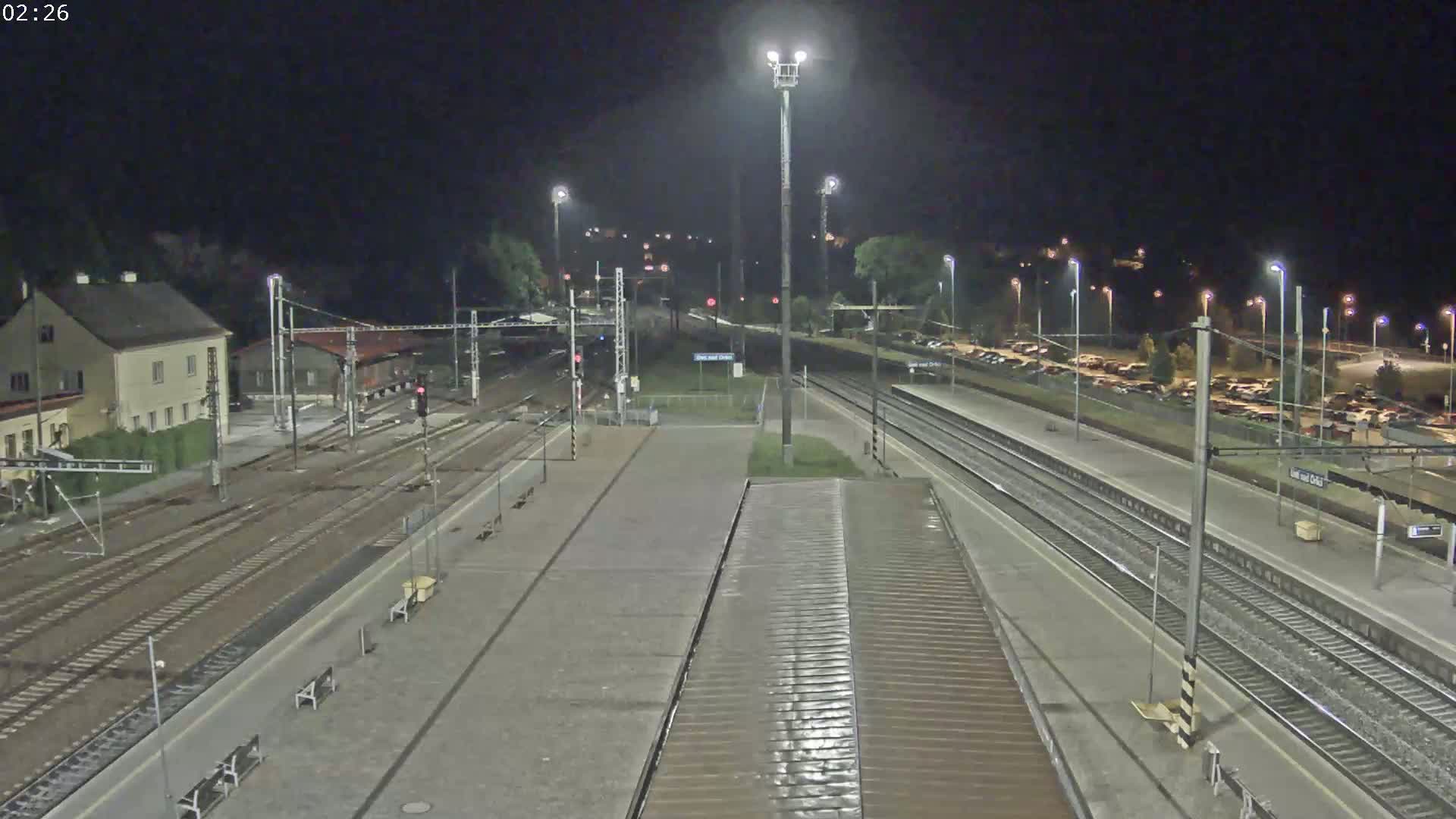 A nighttime view of a train station with multiple tracks, a building, and a parking lot, illuminated by bright lights.