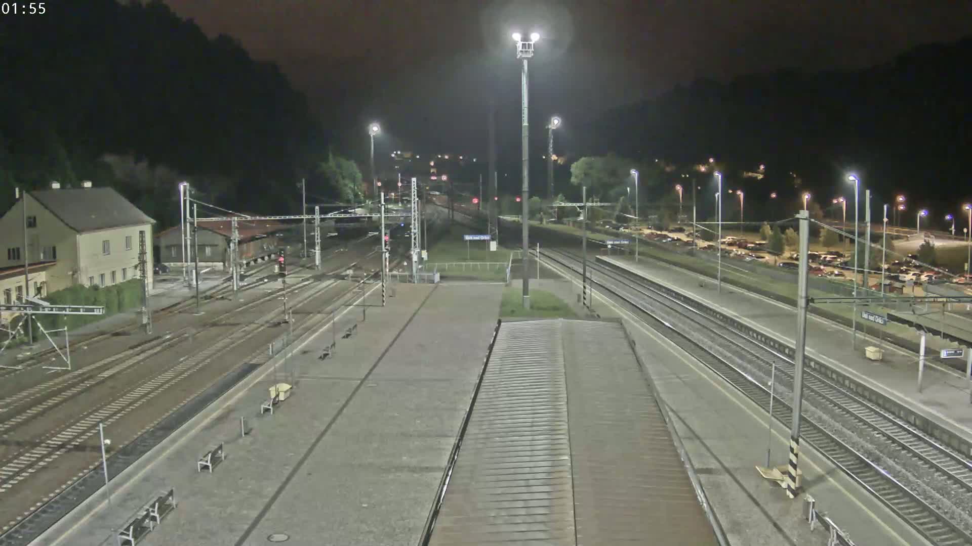 A nighttime view of a train station with multiple tracks, a building, and a parking lot, under clear skies.