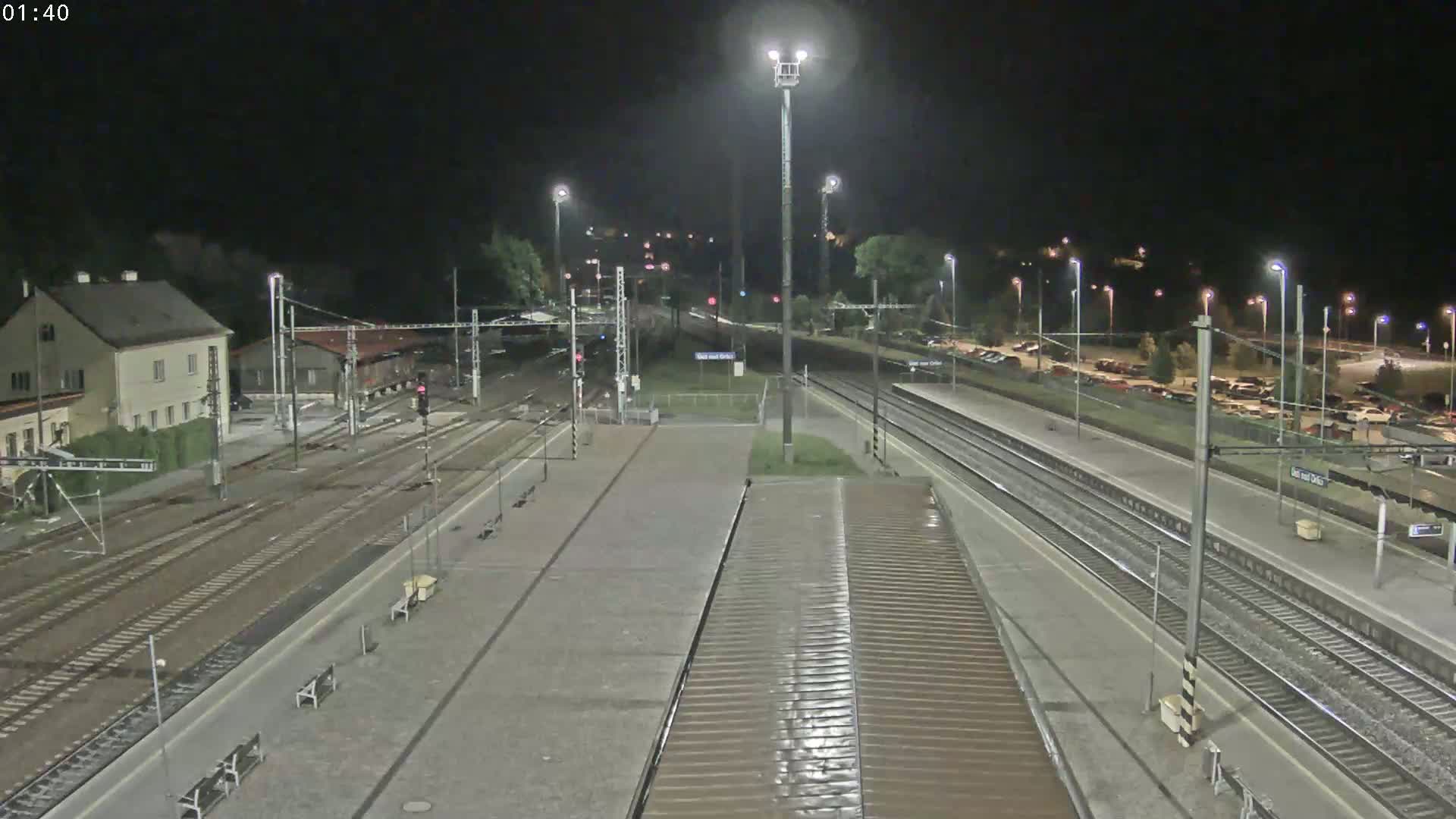 A nighttime, high-angle view of a train station with multiple tracks, platforms, and a parking lot filled with cars.