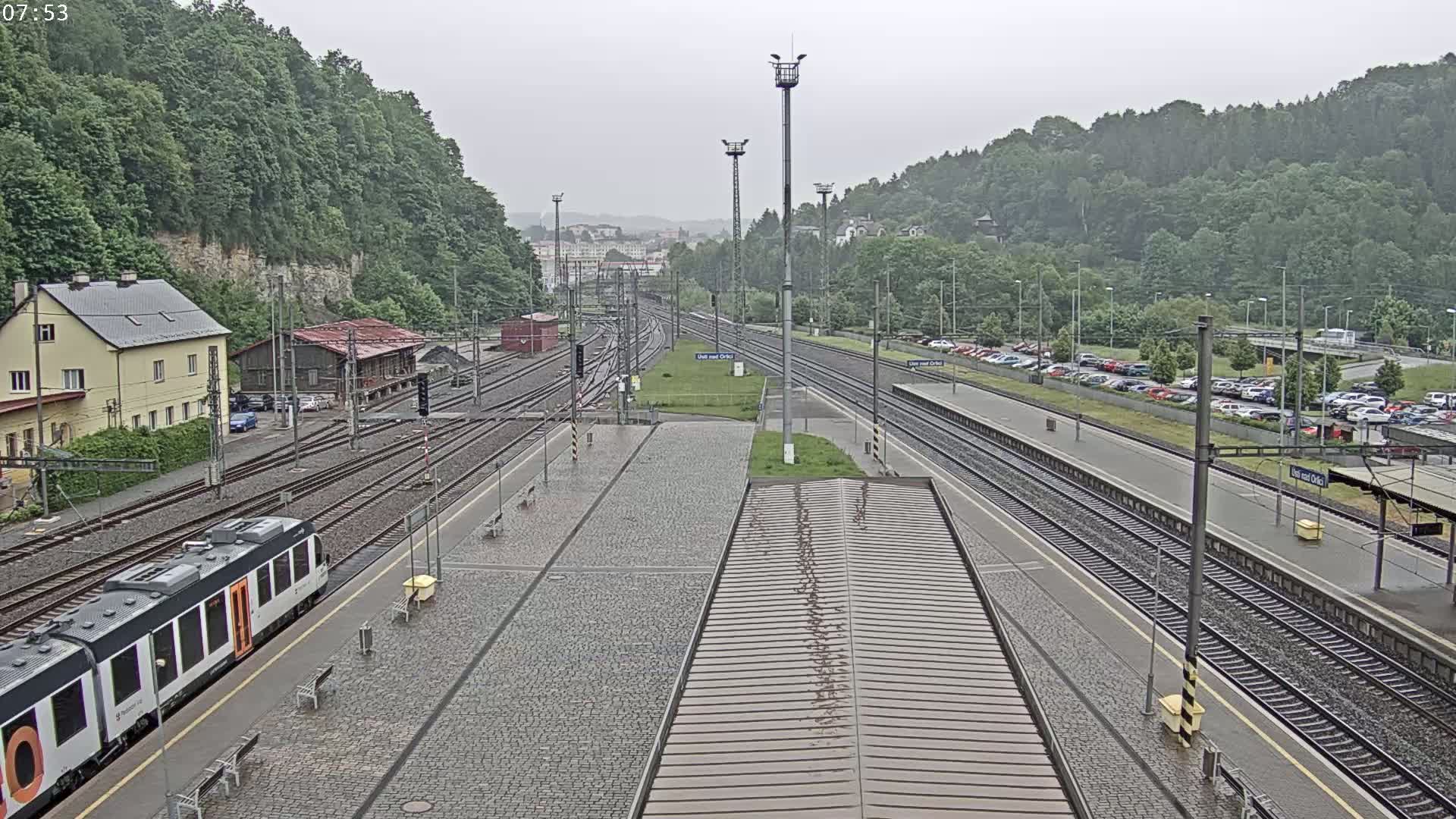 A train sits at a station platform next to multiple train tracks, surrounded by buildings and lush green hills under an overcast sky.